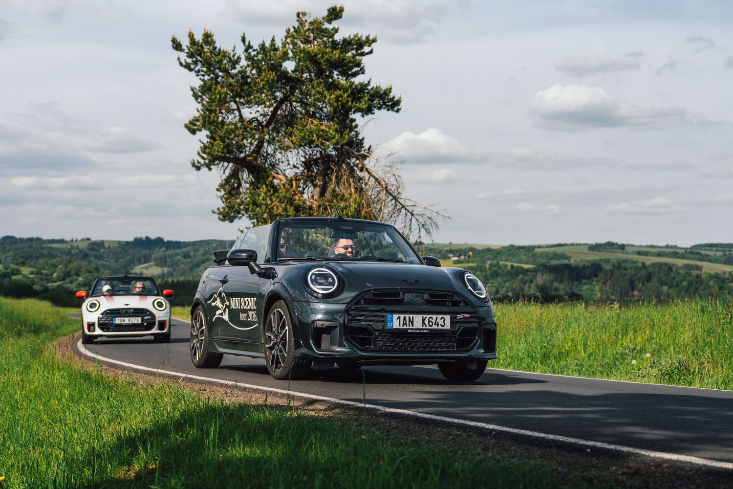 Two MINI Cooper convertibles driving side by side on a scenic rural road with green fields.