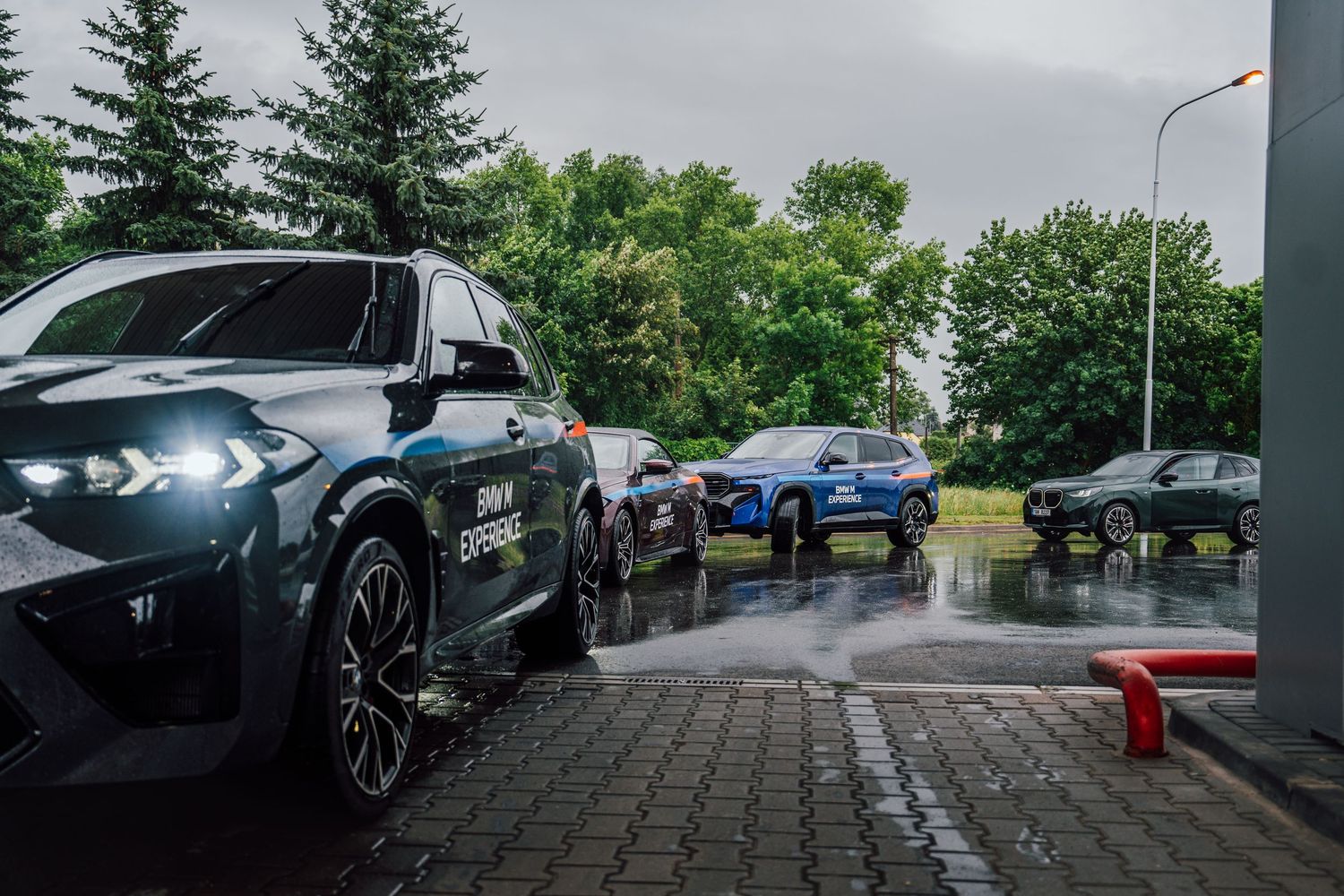 BMW M Experience cars lined up on wet brick surface with headlights on during rainy weather.