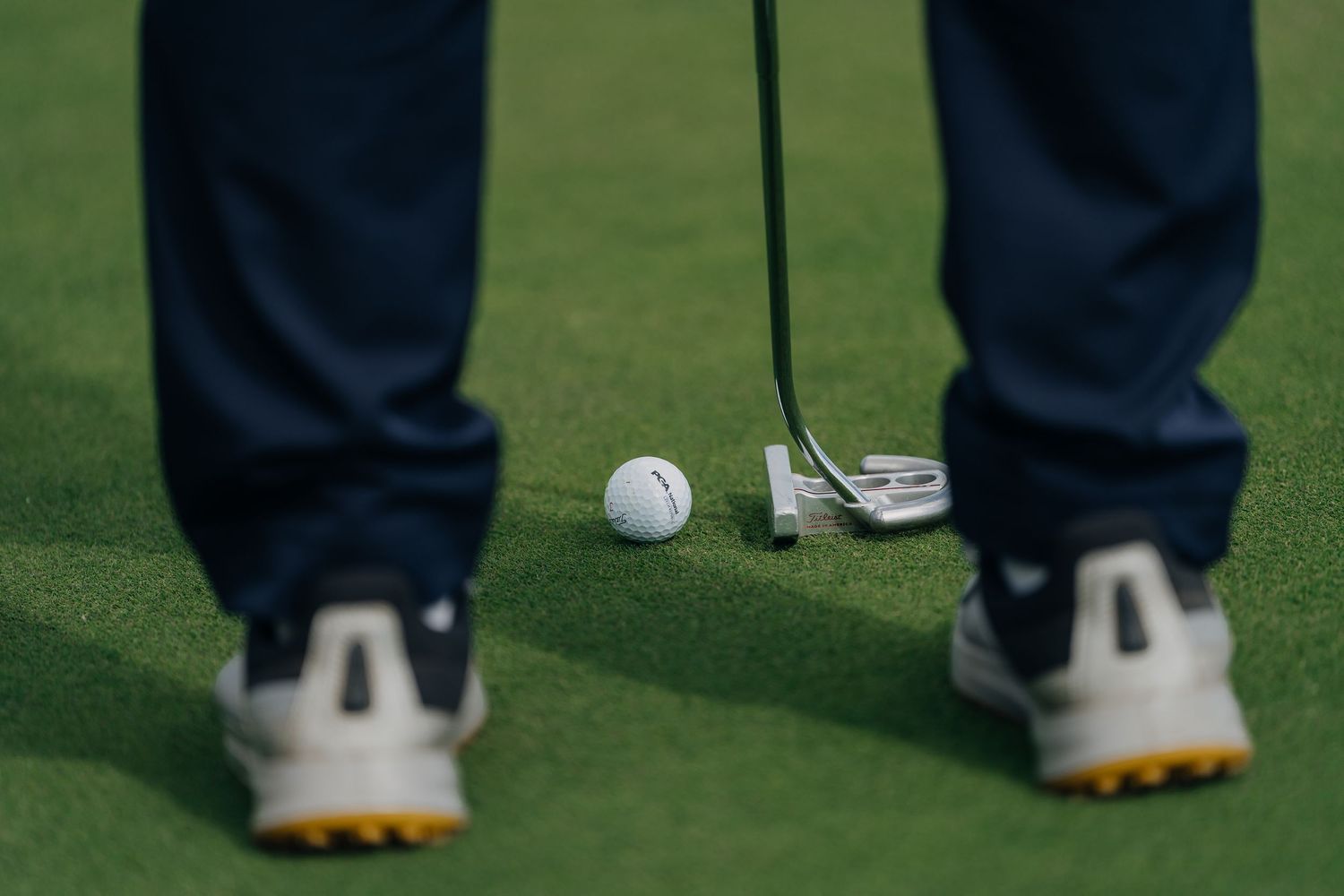 Close-up of a golfer putting on a green with a white golf ball and iron club near white golf shoes.