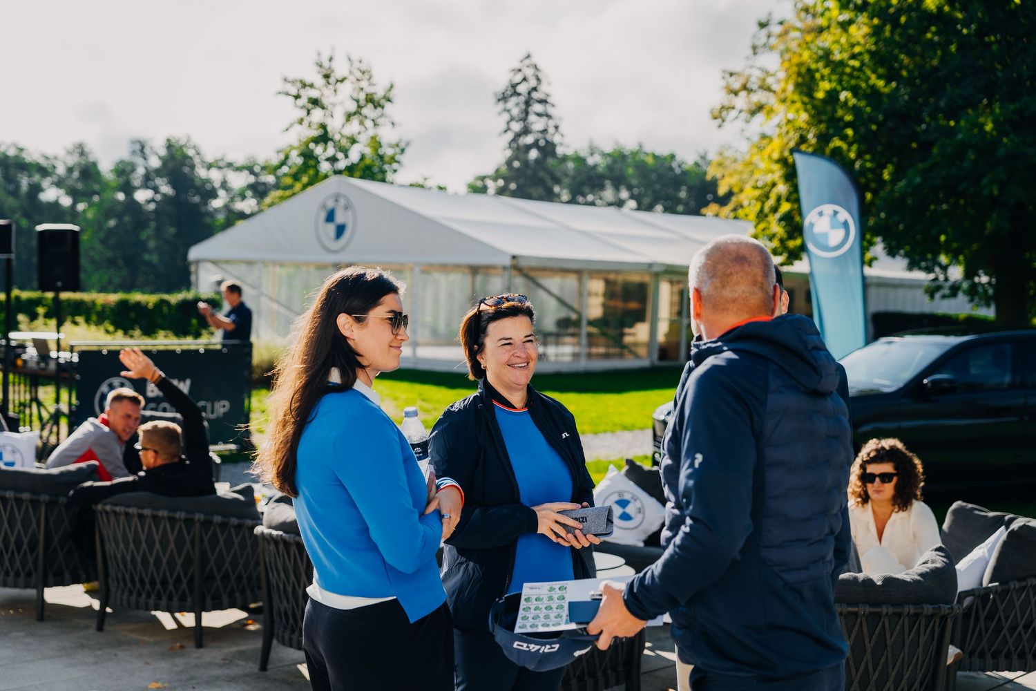 Three people in blue jackets networking outdoors at a BMW golf event on a sunny day.