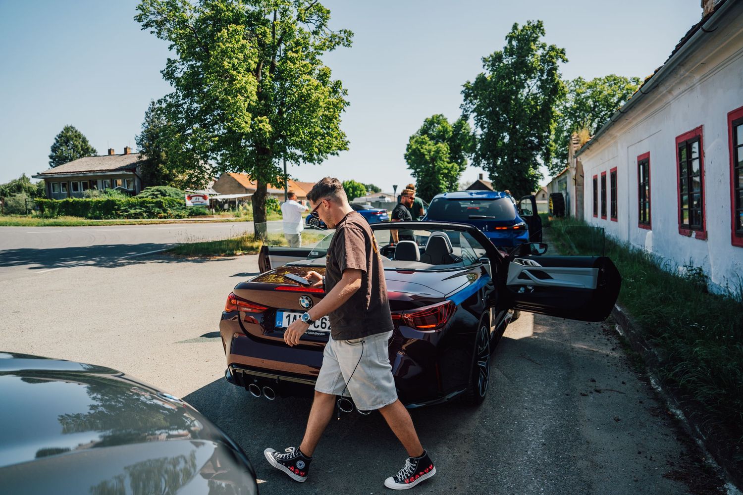 Man walking beside a dark BMW M convertible parked in a sunny village square.