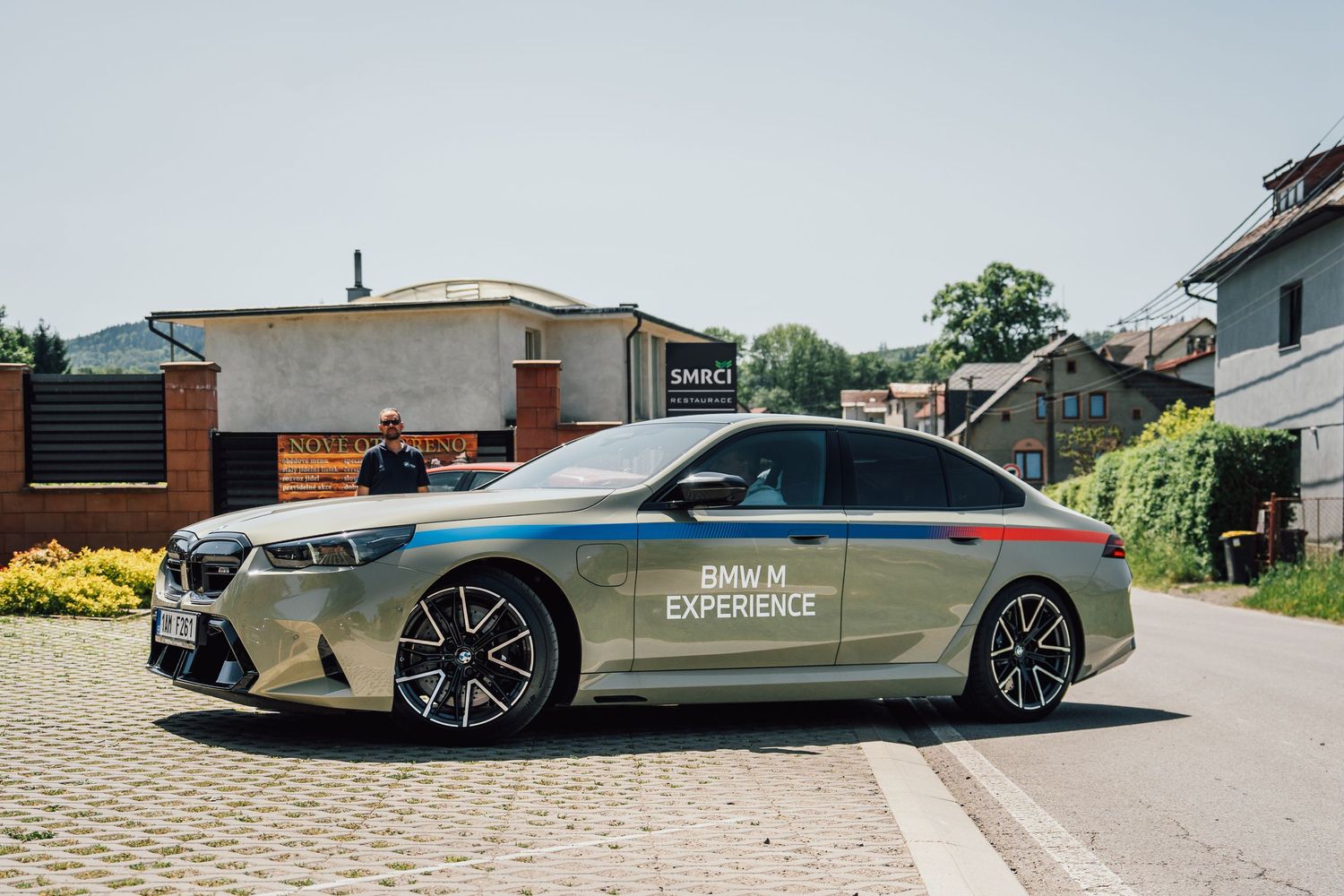 BMW M5 with M Experience livery parked in a village square on a bright sunny day.