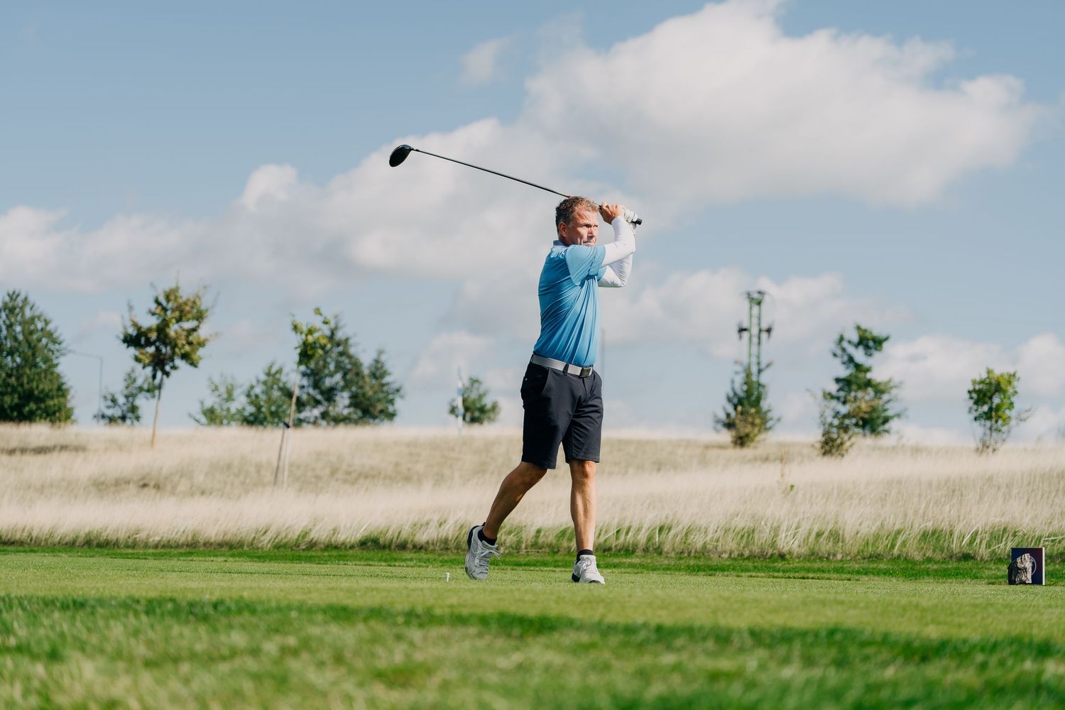 Golfer in blue shirt completing a drive swing on a sunny golf course with green fairway and trees.