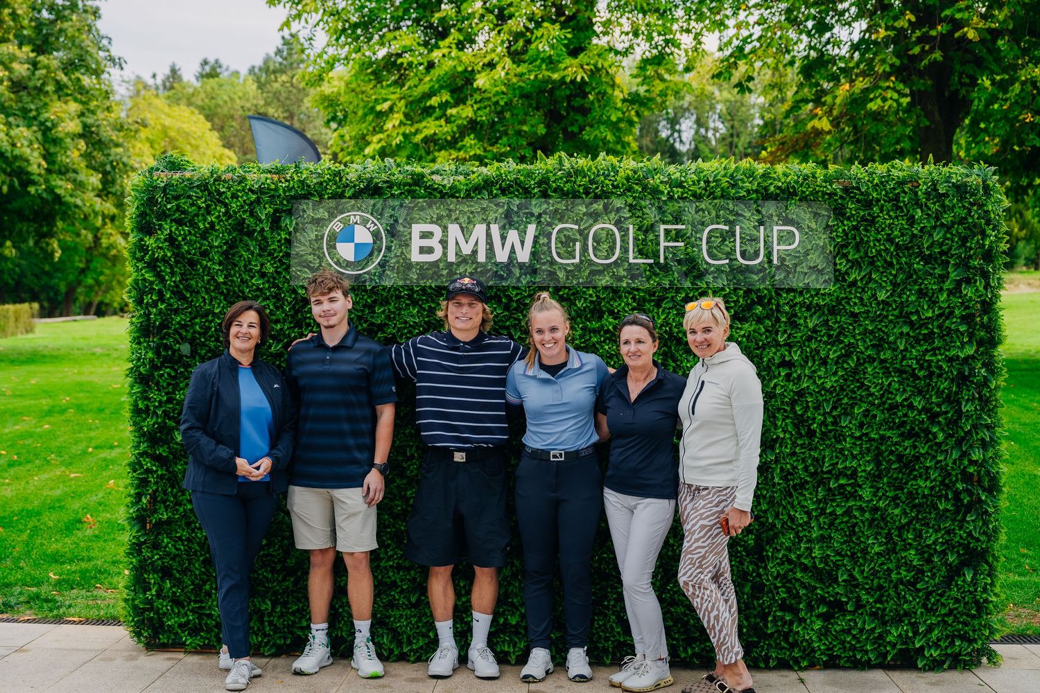 Group of six golfers posing together in front of a BMW Golf Cup hedge sign at a golf event.
