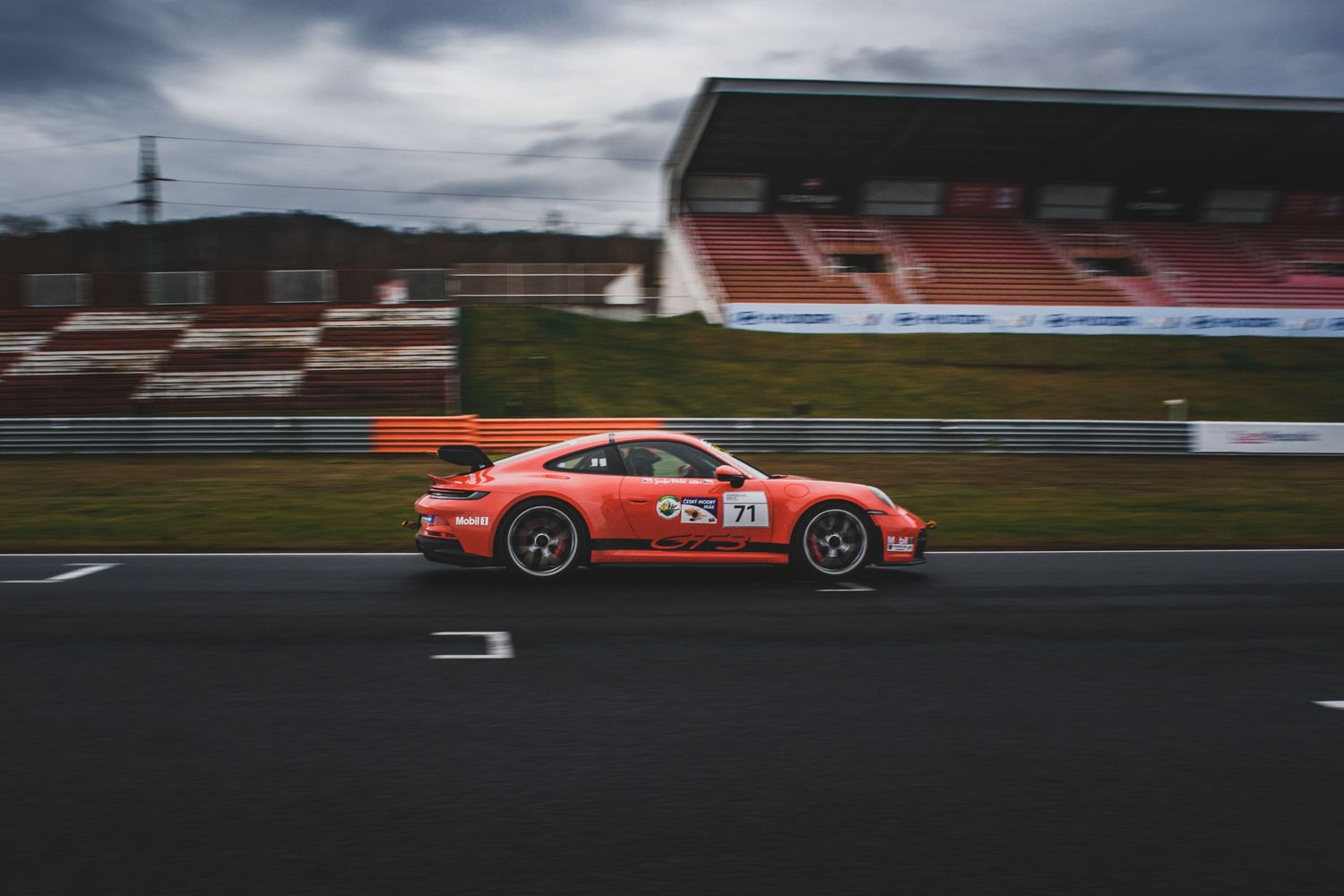 Orange Porsche 911 GT3 Cup race car number 37 racing on a track with grandstands behind.