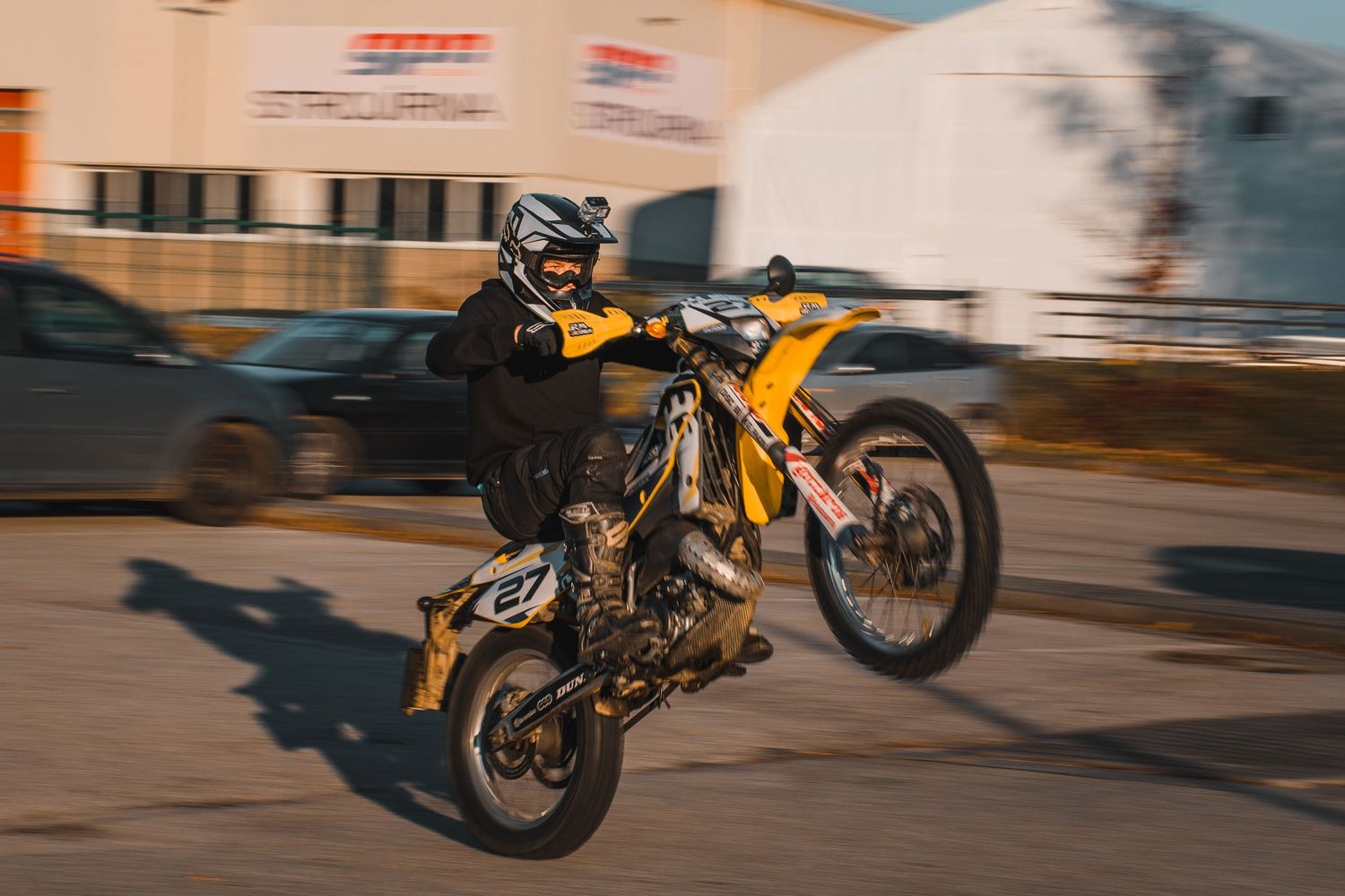 Rider performing wheelie on yellow dirt bike in industrial parking area during golden hour.
