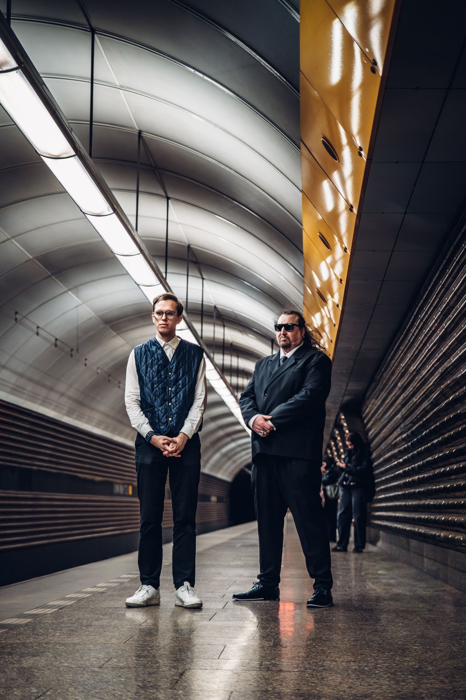 Two men standing on modern subway platform, one in casual wear and one in black suit with sunglasses.