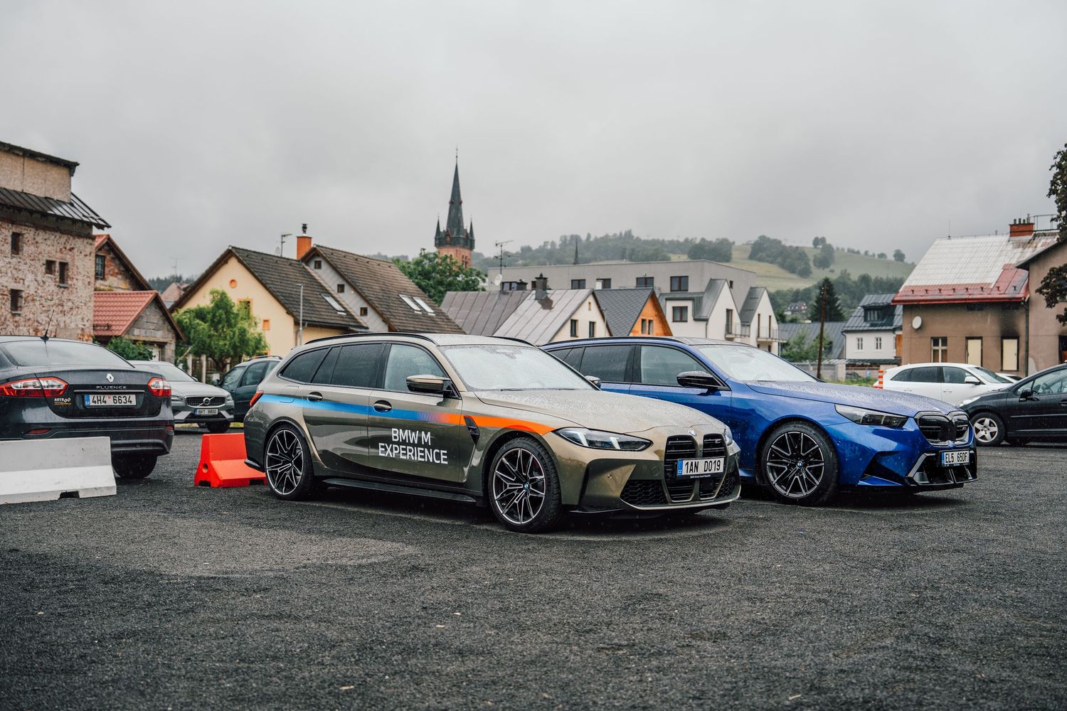 Two BMW M cars parked in a European village, including an M3 Touring with BMW Experience livery and a blue BMW M car.