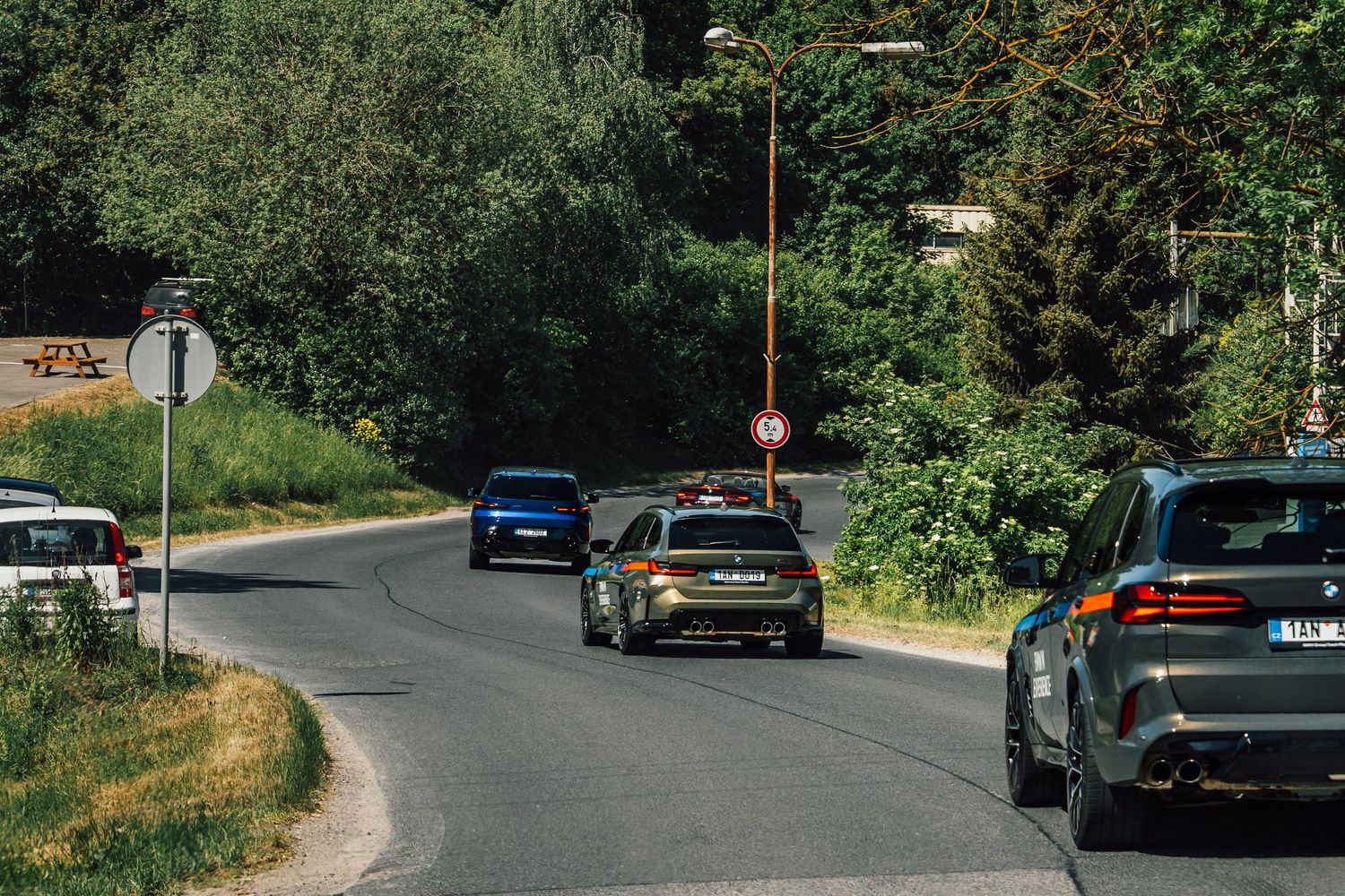 Multiple BMW vehicles navigating a winding road through a lush green forested area.