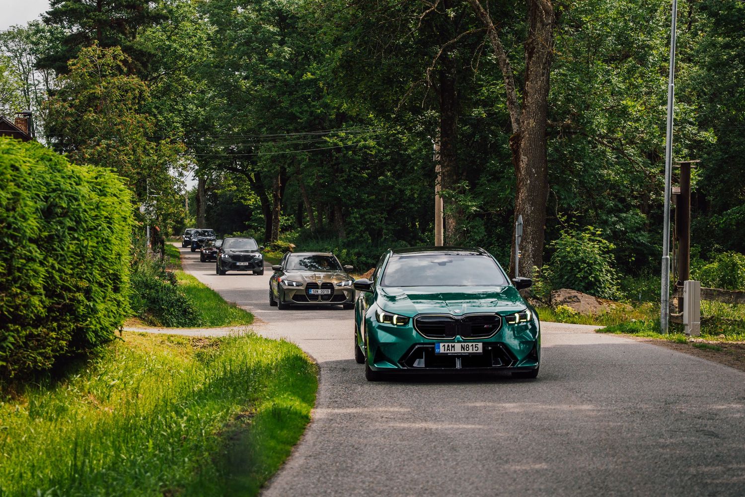 Green BMW M3 leads a convoy of cars along a tree-lined country road on a scenic driving tour.