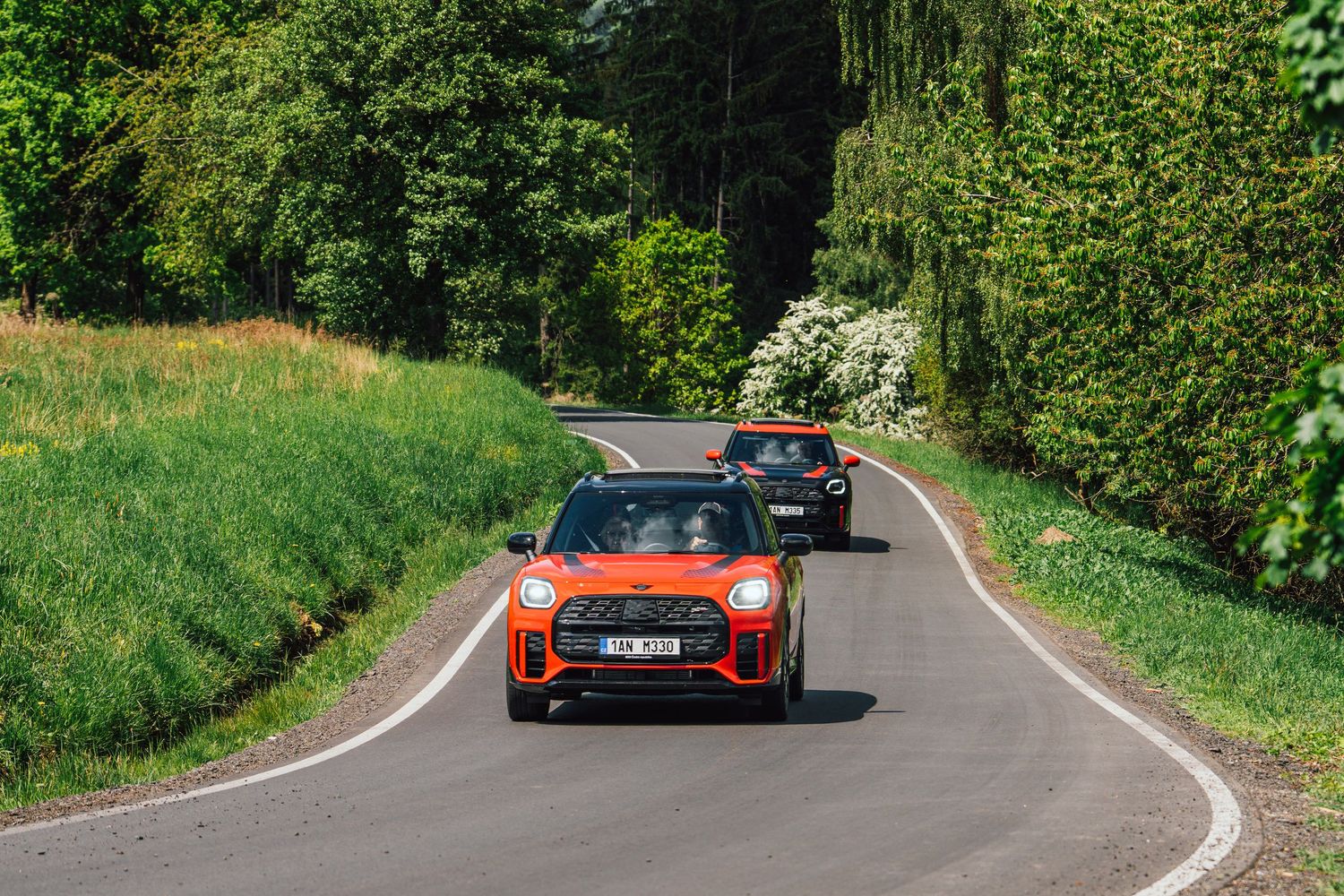 Two orange MINI Cooper cars driving along a winding tree-lined country road on a sunny day.