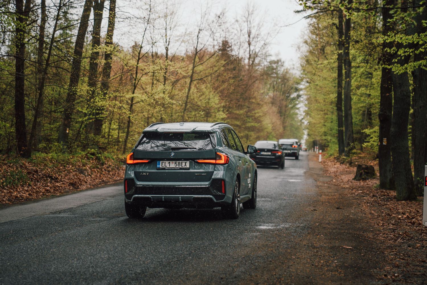 Rear view of a dark green BMW iX1 driving on a wet forest road flanked by autumn trees.