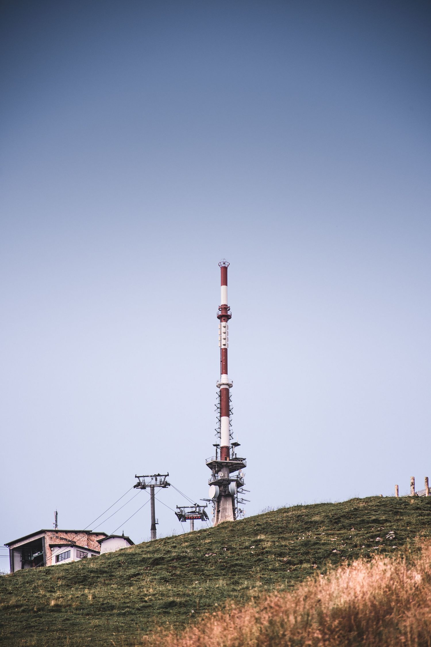 Tall red and white radio transmission tower on green hillside against clear blue sky.