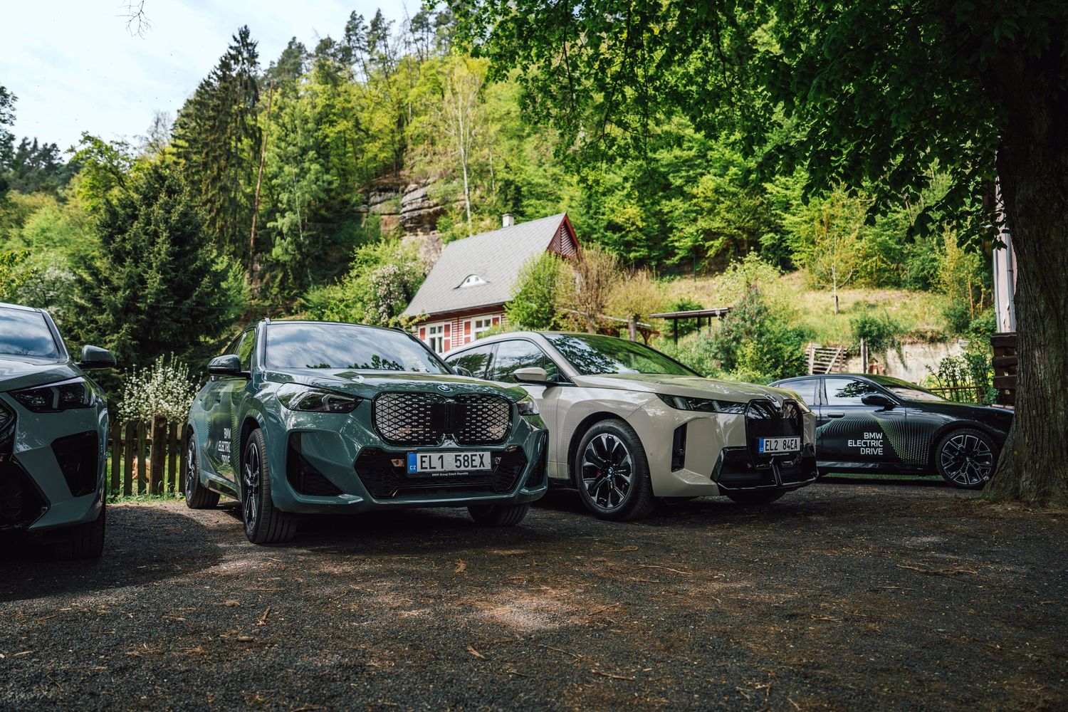 Multiple BMW electric SUVs parked in a wooded area near a rustic cottage during a BMW Electric Drive event.