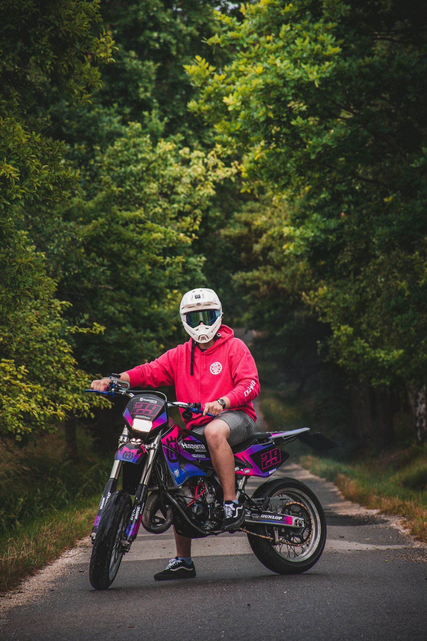 Motocross rider wearing red hoodie and white helmet poses on colorful purple dirt bike on forest road.
