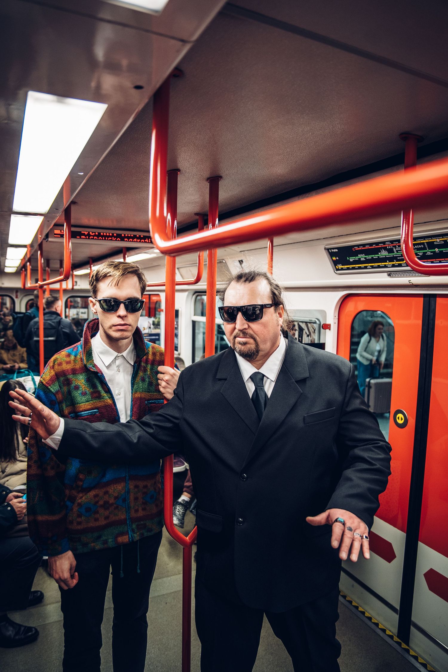 Two men in sunglasses posing inside subway car, one in colorful jacket and one in black suit.