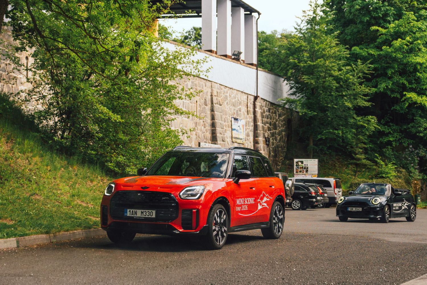 Red MINI Cooper JCW leads a group of MINI cars on a tree-lined road near a stone building.