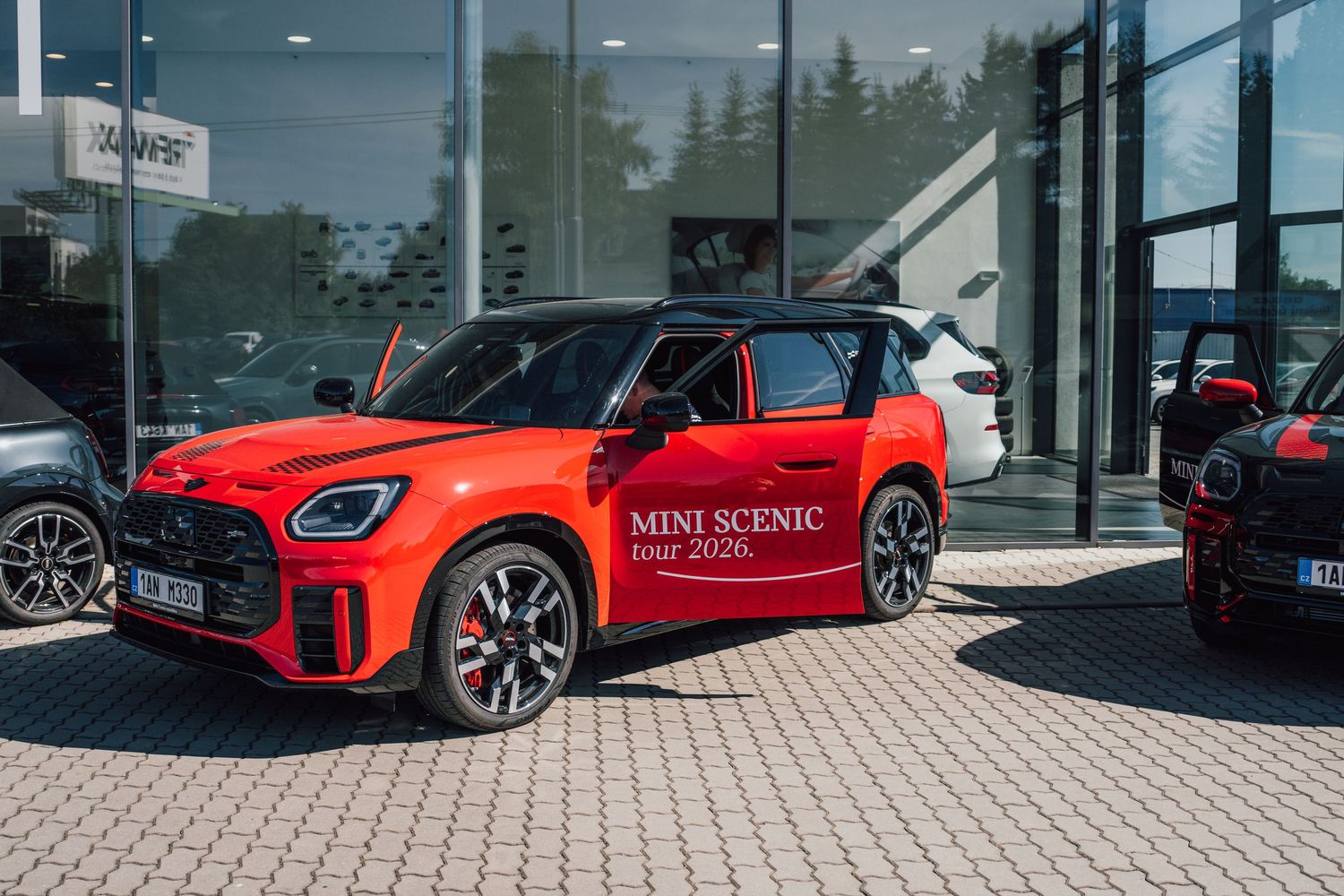 Red MINI Scenic tour 2026 SUV displayed outside modern glass BMW dealership building.