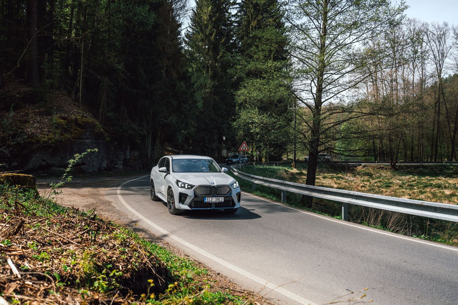 White BMW SUV driving on a scenic forest road with guardrails and tall trees in early spring.