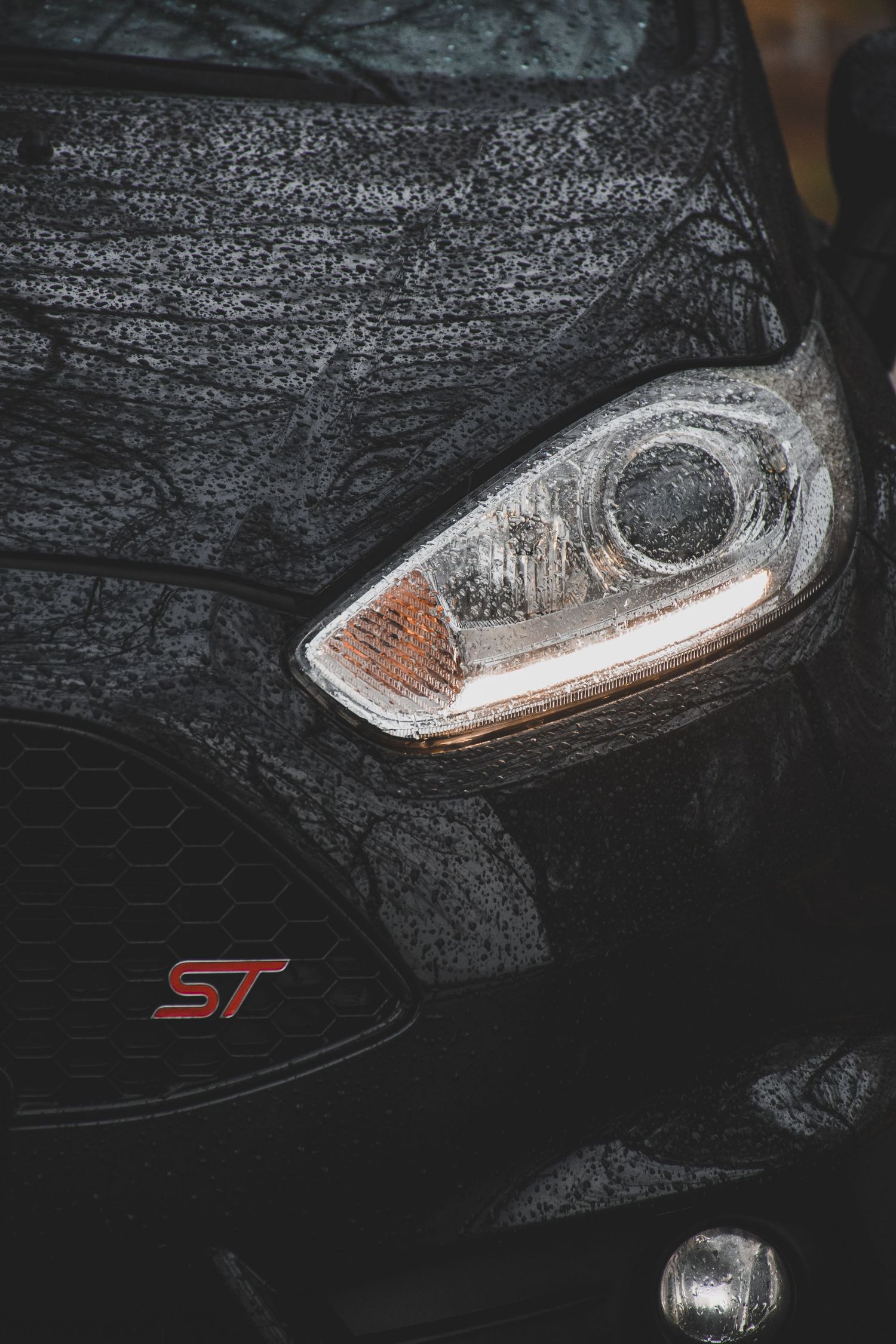 Black Ford Fiesta ST covered in rain droplets with illuminated headlight in dark conditions.