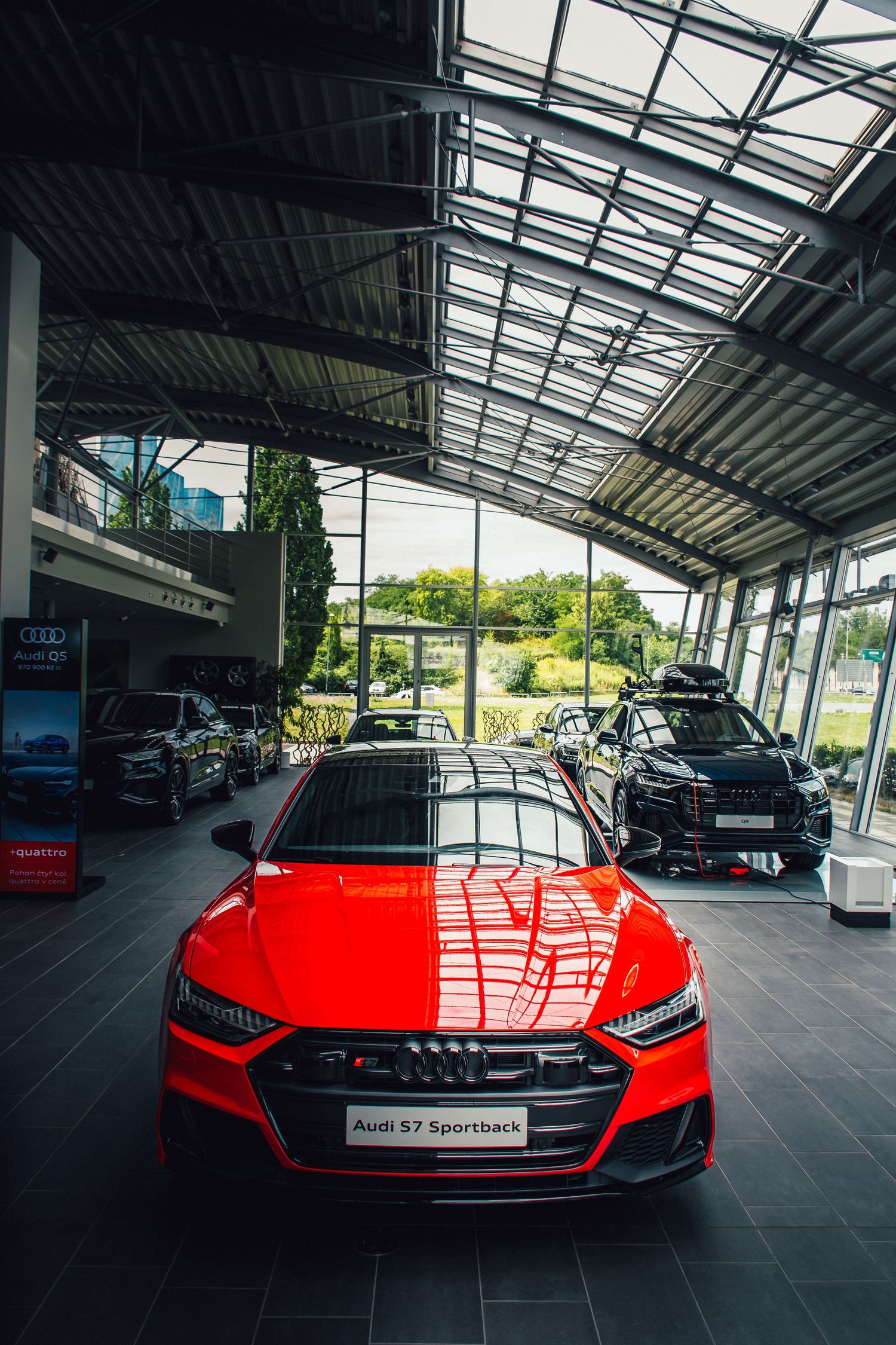 Red Audi S7 Sportback on display in a bright modern dealership showroom with glass ceiling and multiple vehicles.