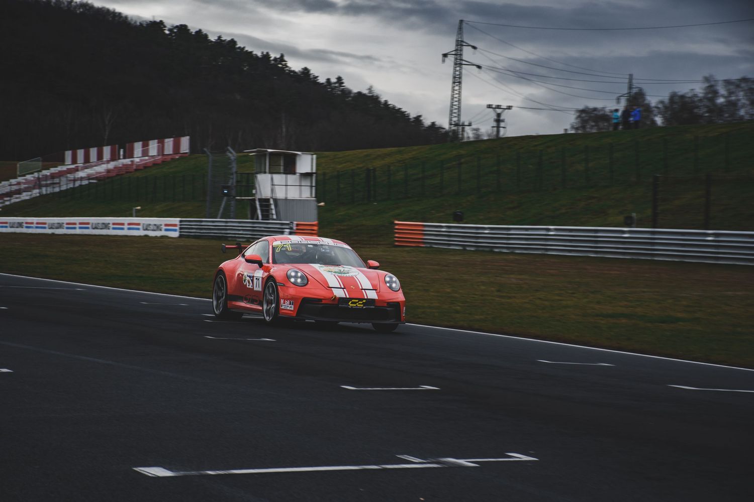 Orange Porsche 911 GT3 race car on a wet track circuit under cloudy skies.
