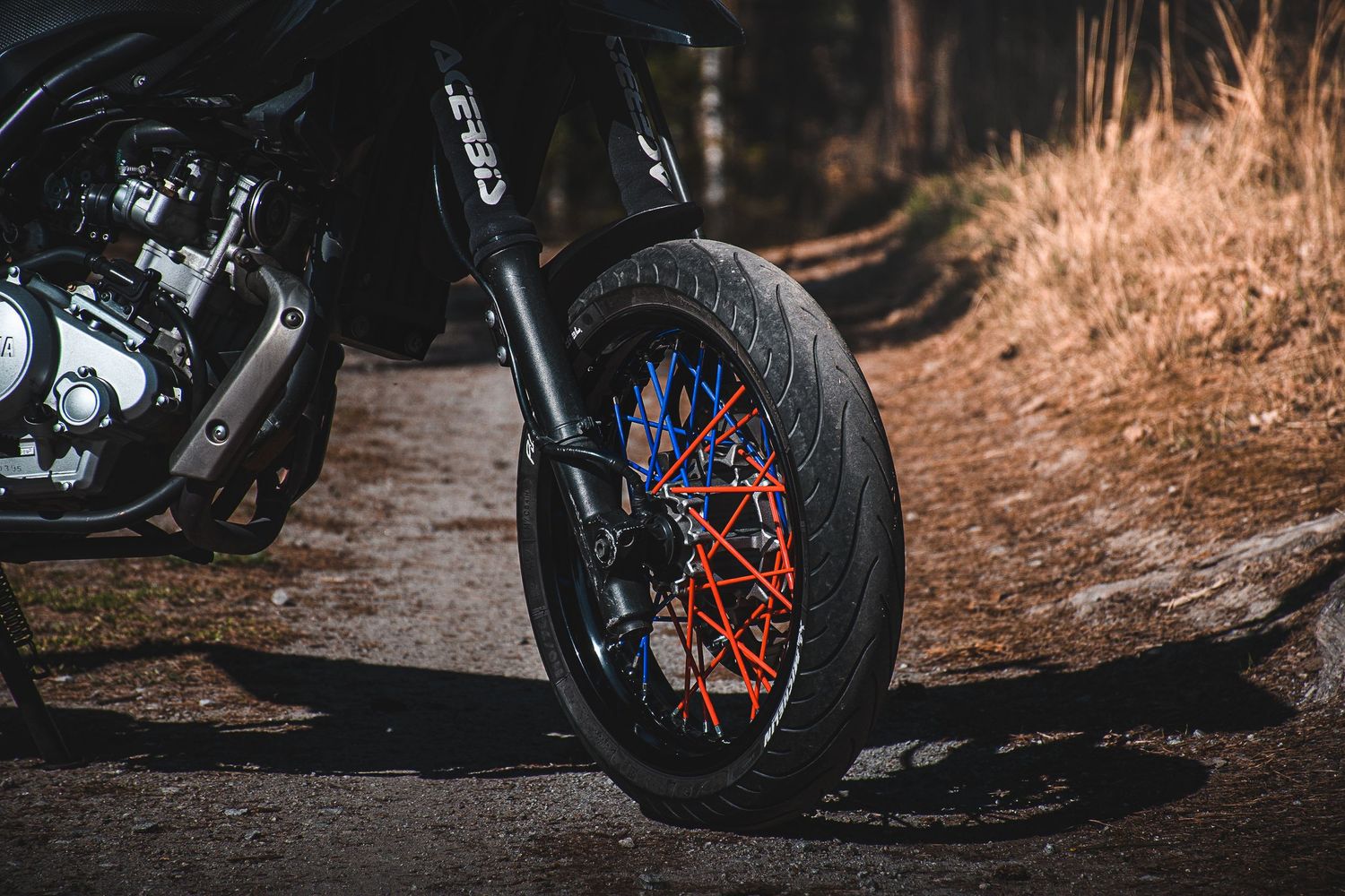 Black supermoto motorcycle front wheel with colorful blue and red spokes on a forest trail.