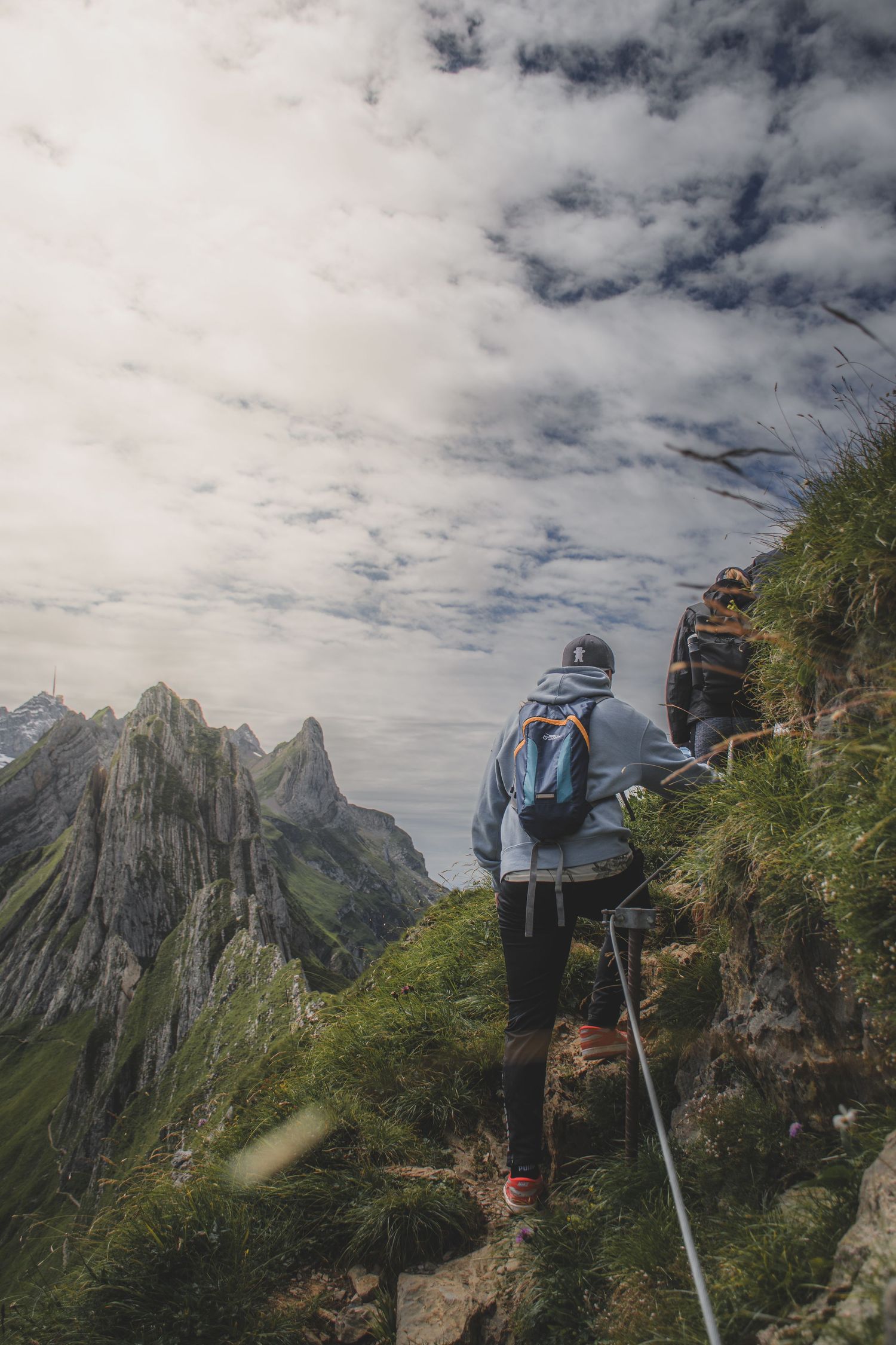 Hikers with backpacks descend a steep mountain trail secured by a rope with dramatic rocky peaks behind.