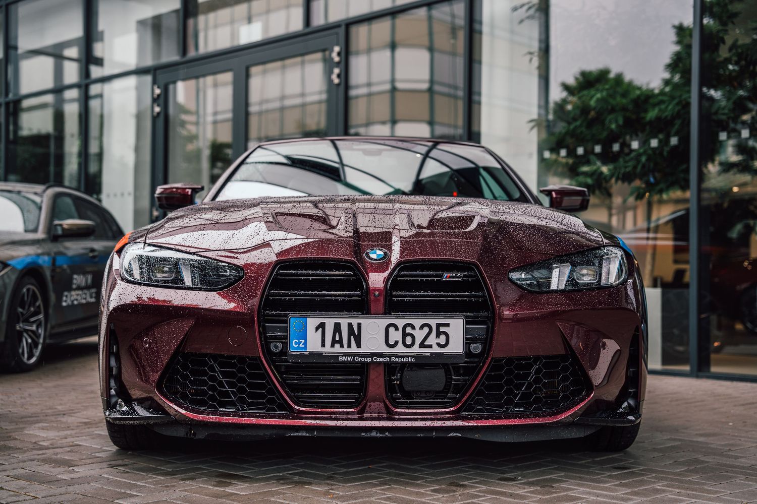 Front view of a dark red BMW M3 with Czech license plate parked outside a modern glass building.