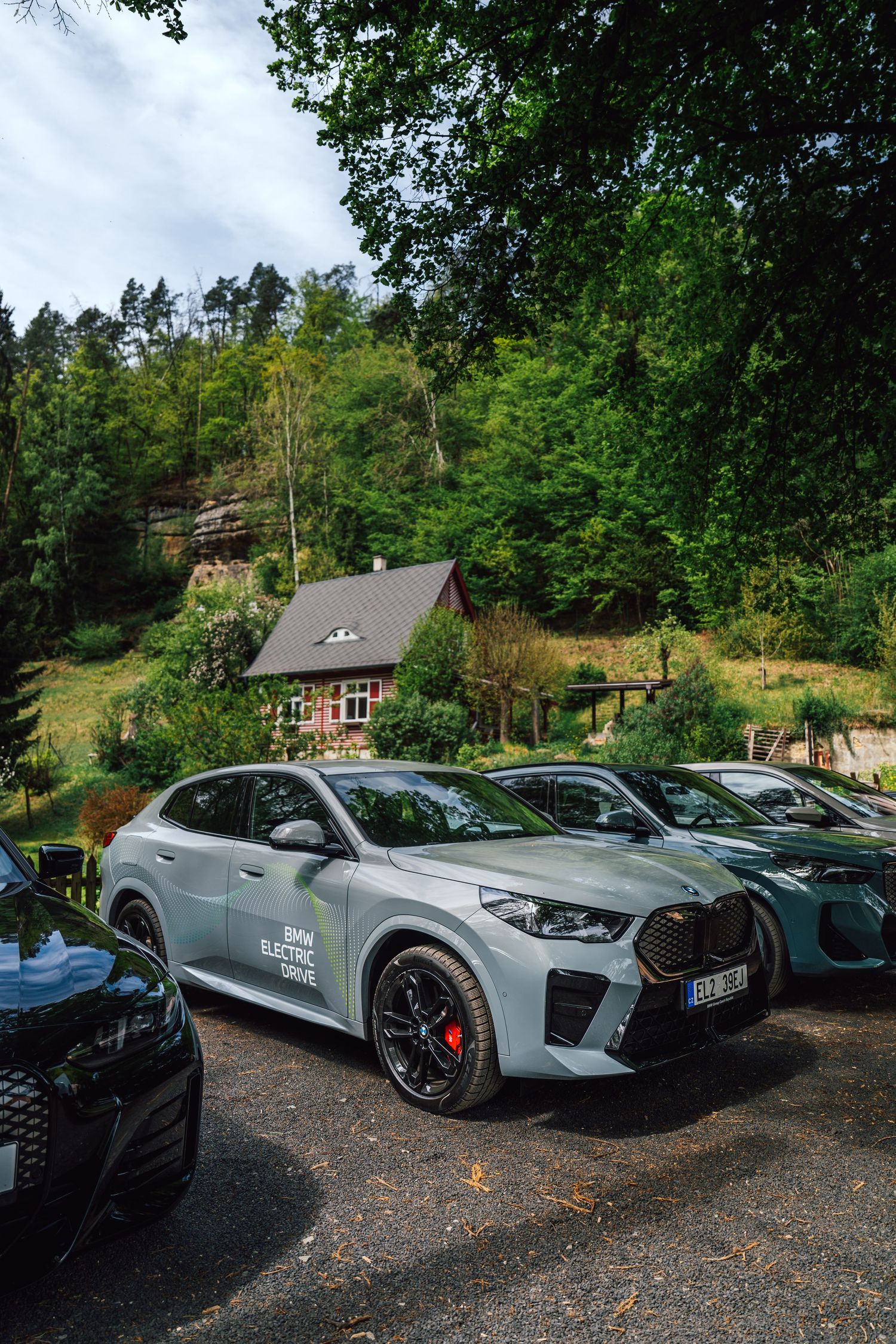 Gray BMW iX M60 with BMW Electric Lodge branding parked in a forest clearing near a hillside cottage.