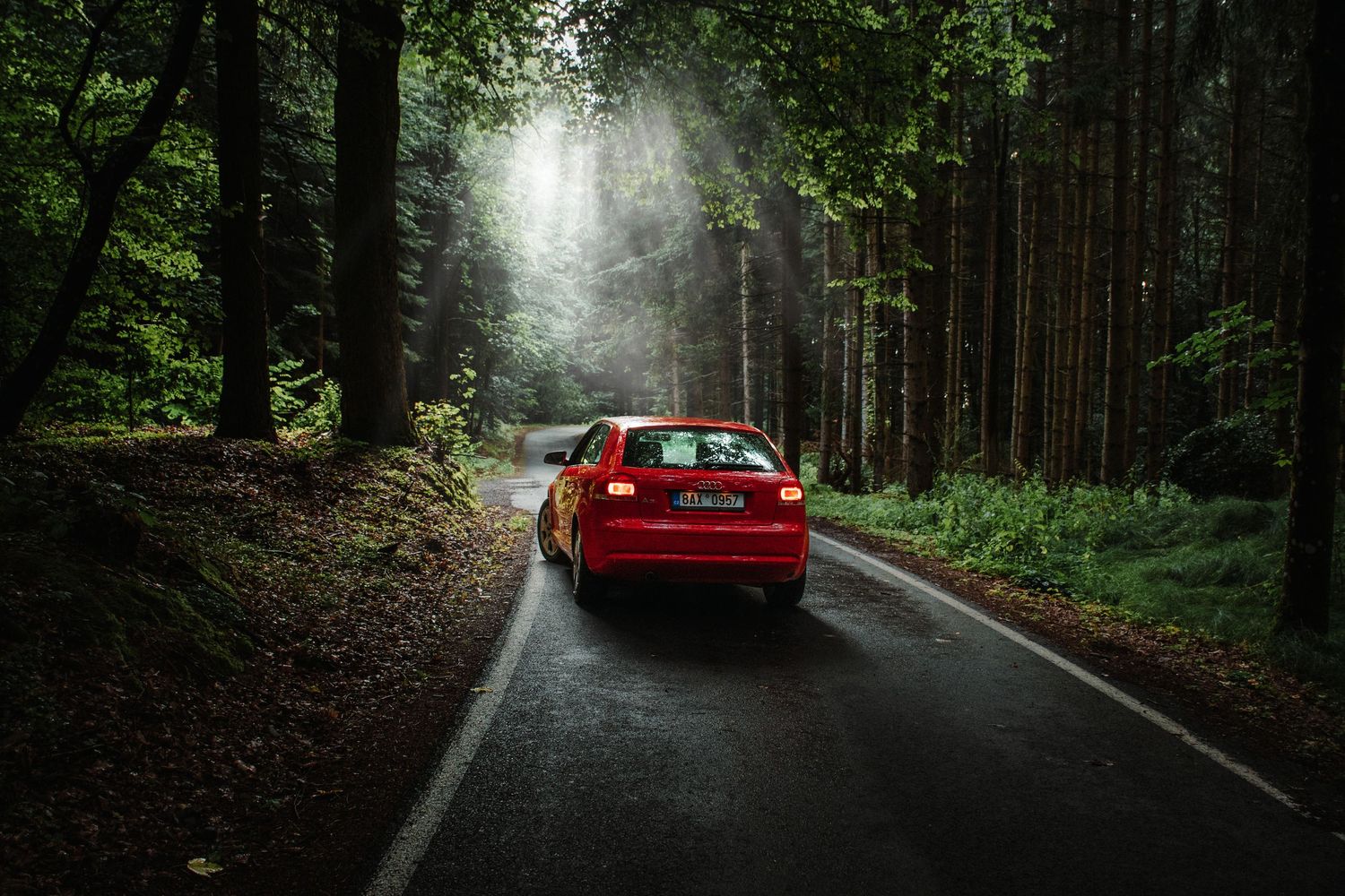 Red Audi sedan driving through a misty forest road with sunlight filtering through trees.