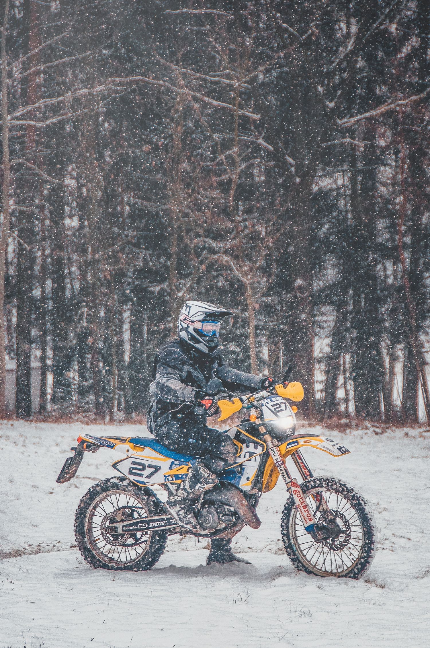 Dirt bike rider number 27 riding a yellow Husqvarna through heavy snowfall in a winter forest.