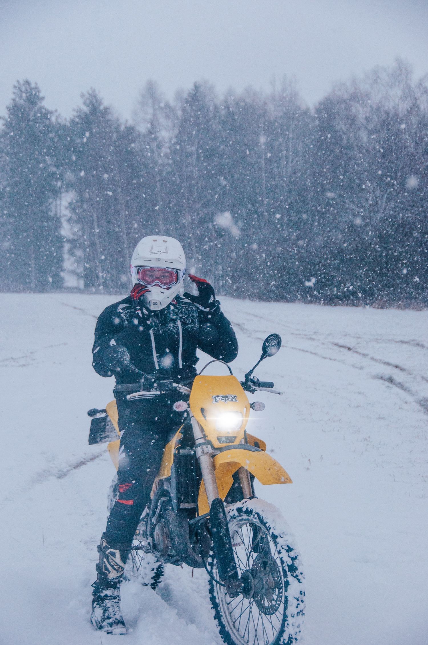 Motocross rider in white helmet adjusts gear on yellow dirt bike during heavy blizzard.