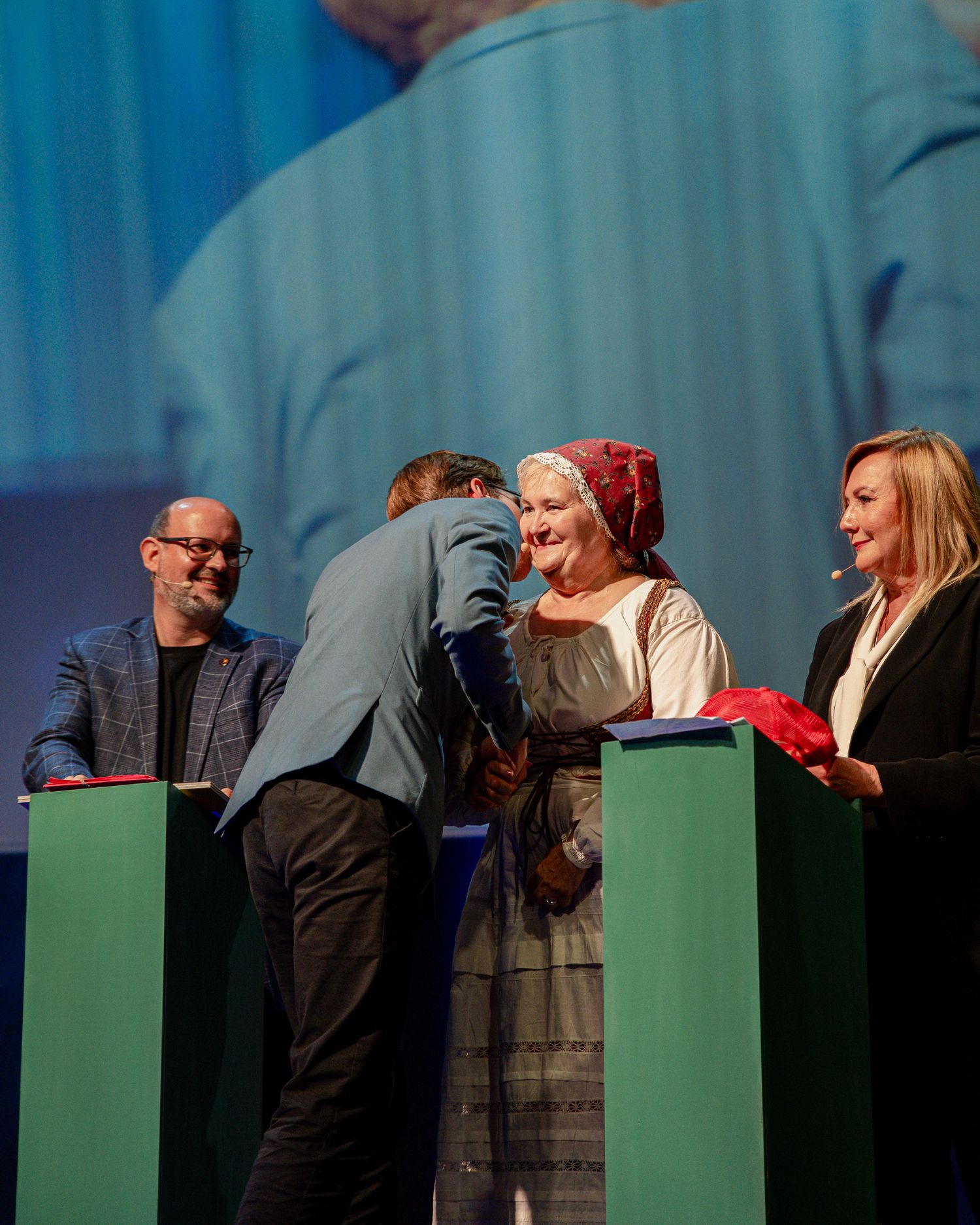Man in blue suit leaning to greet elderly woman in traditional dress on stage during ceremony.