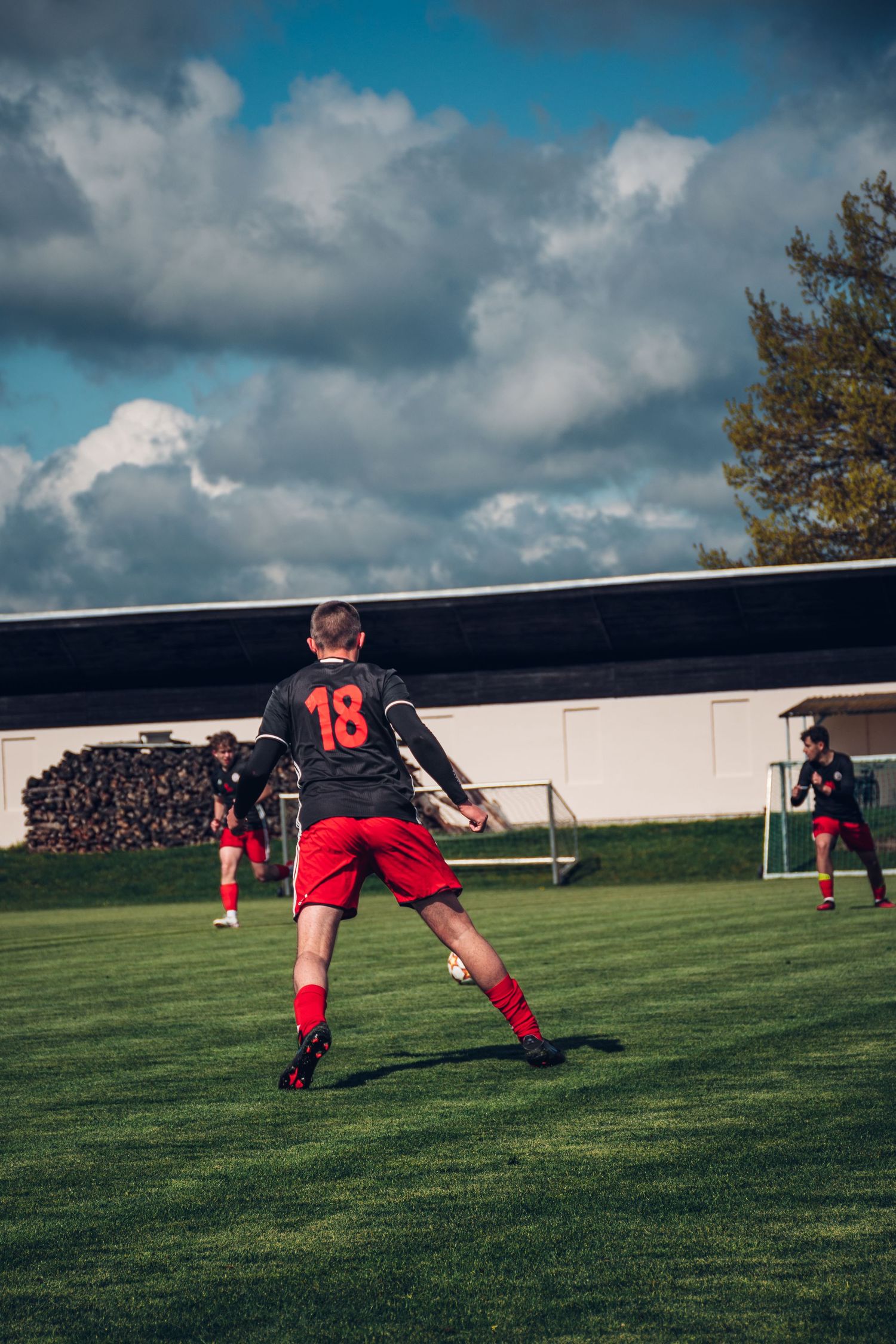 Soccer player number 18 in black and red uniform kicks a ball on a green field under a cloudy blue sky.