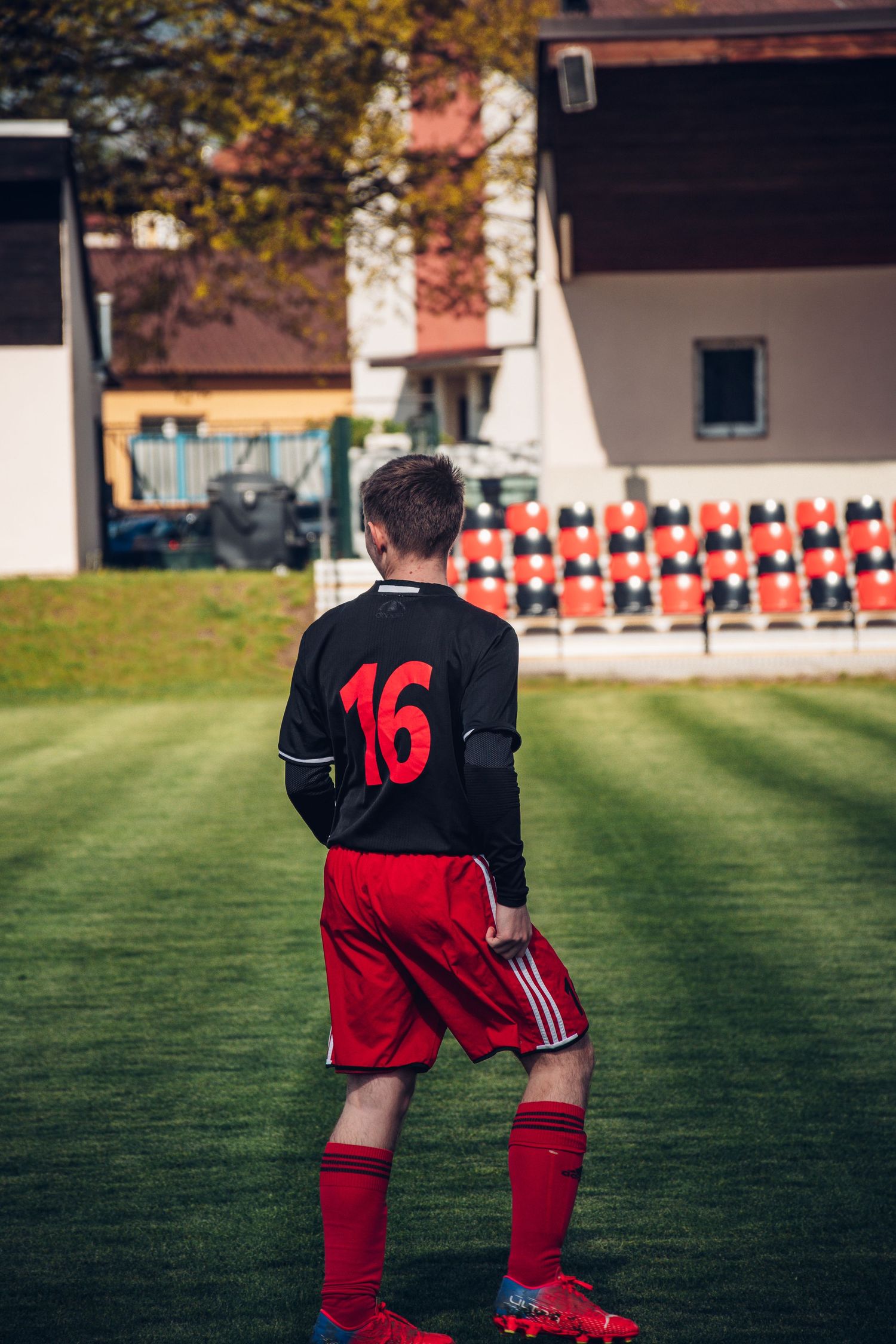 Youth soccer player wearing number 16 black and red uniform stands on a green field with stadium seats behind.