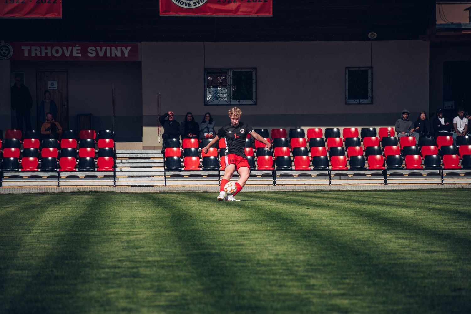 A soccer player dribbles a ball on a green pitch at Trhove Sviny stadium with red empty seats in background.