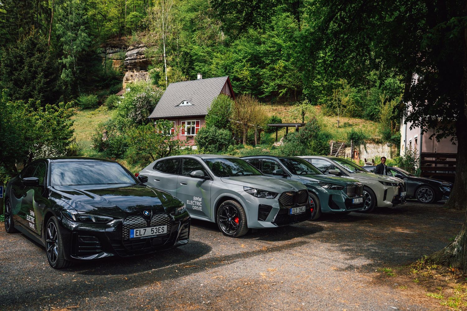 Four BMW electric vehicles parked on a gravel lot in front of a wooded hillside with a red cottage.