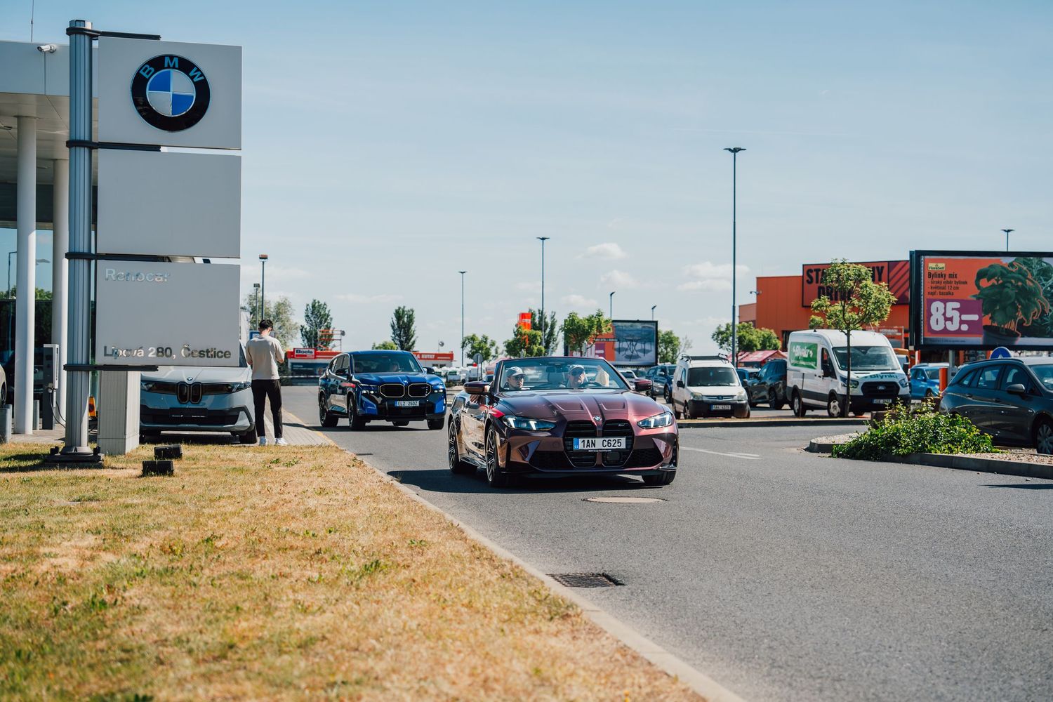 Dark red BMW M4 convertible with M Experience livery driving past a BMW dealership on a busy road.