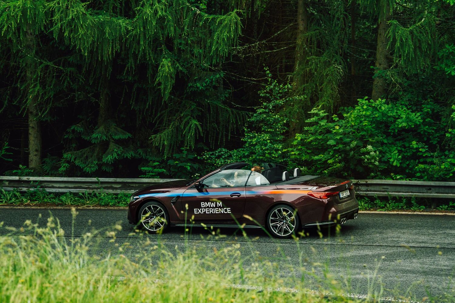 BMW M Experience convertible sports car driving along a lush green forest road in the rain.