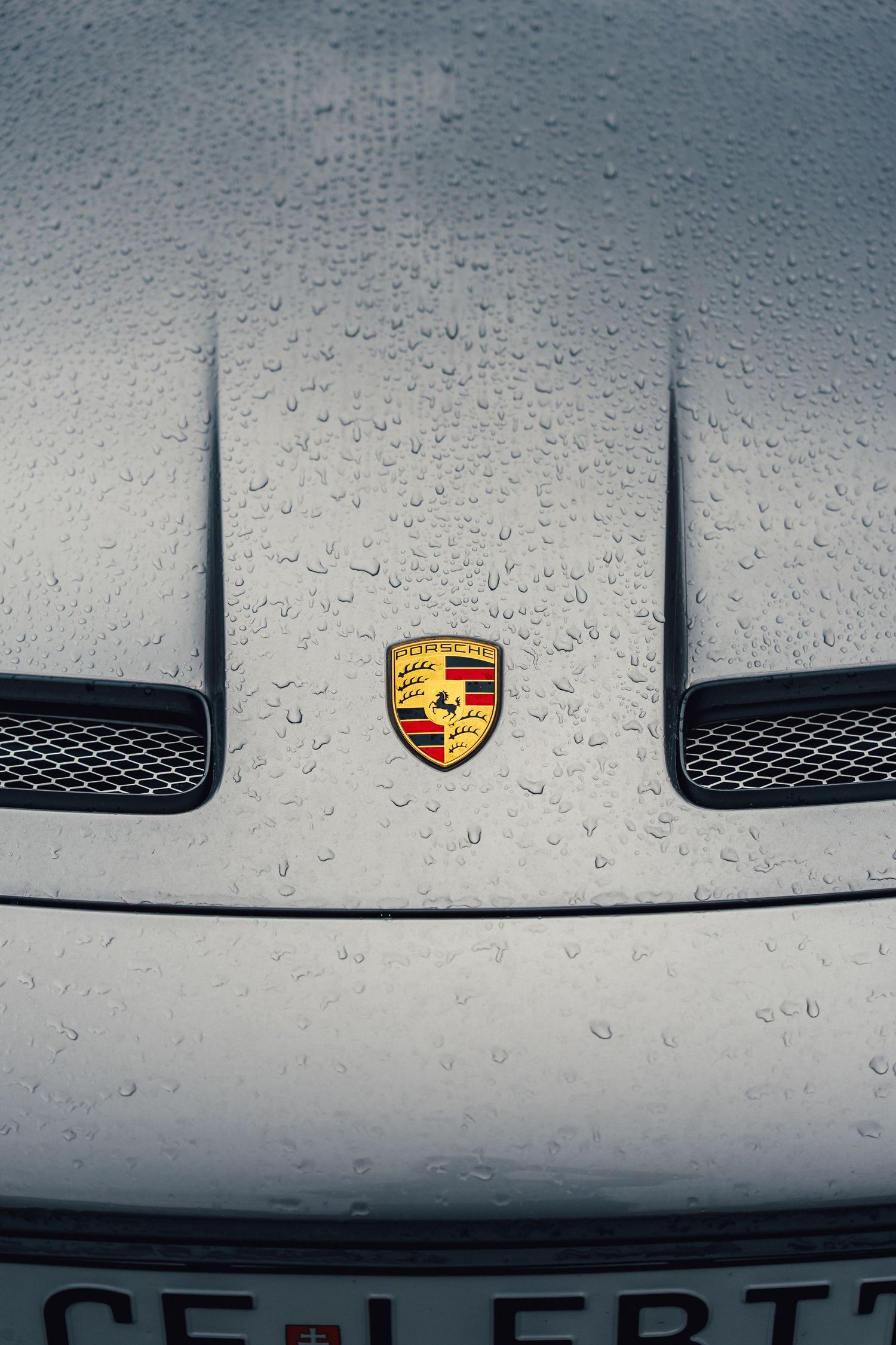 Close-up of a wet Porsche hood with iconic yellow crest emblem covered in rain droplets.
