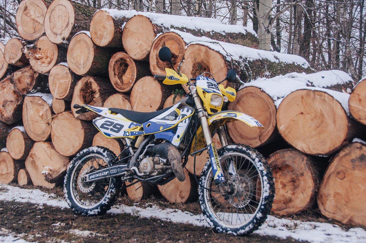 Yellow Husqvarna number 25 dirt bike parked against stack of cut logs in winter snow.