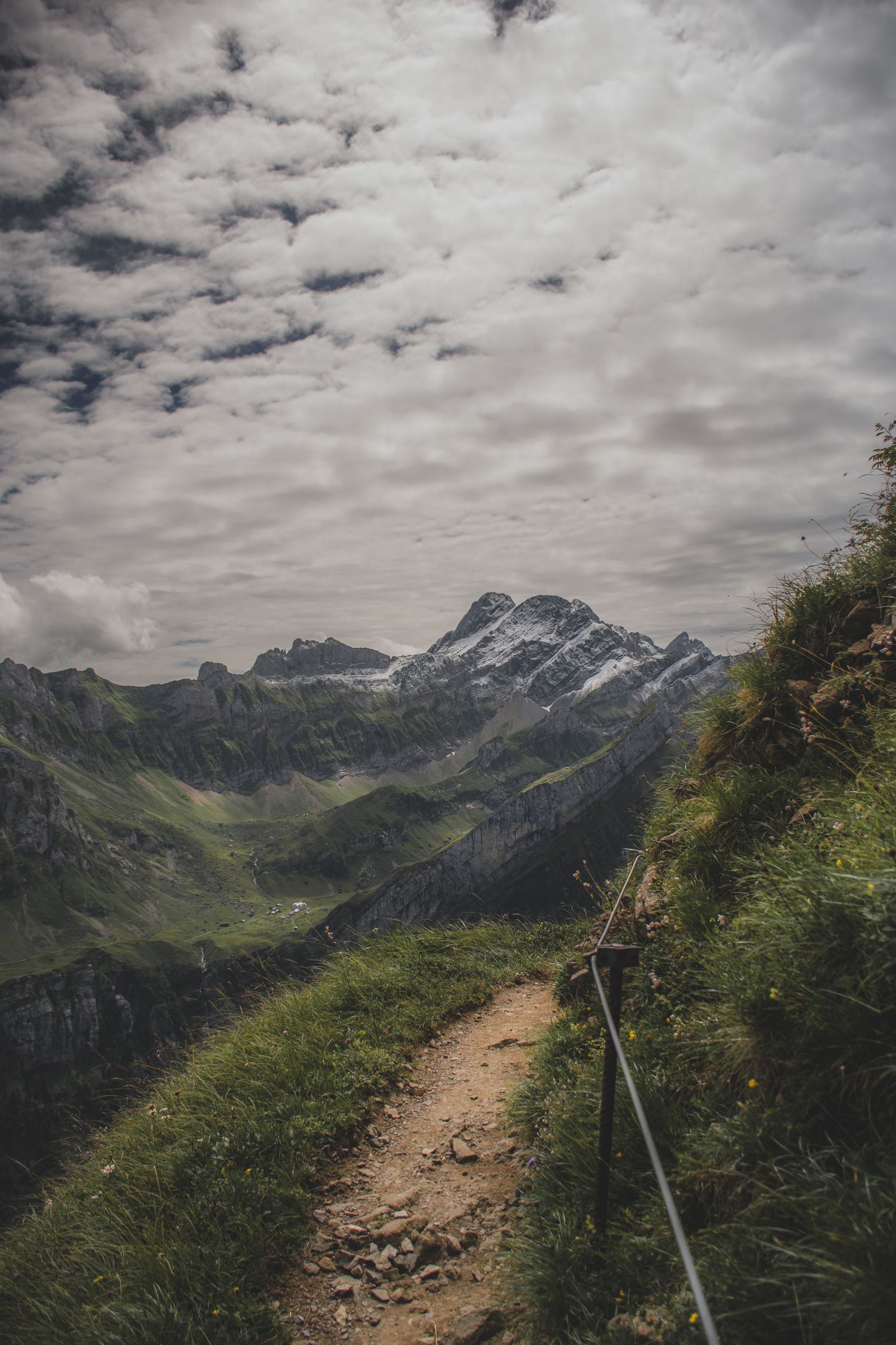 A narrow hiking trail winds along a steep mountain slope toward snow-capped peaks under cloudy skies.