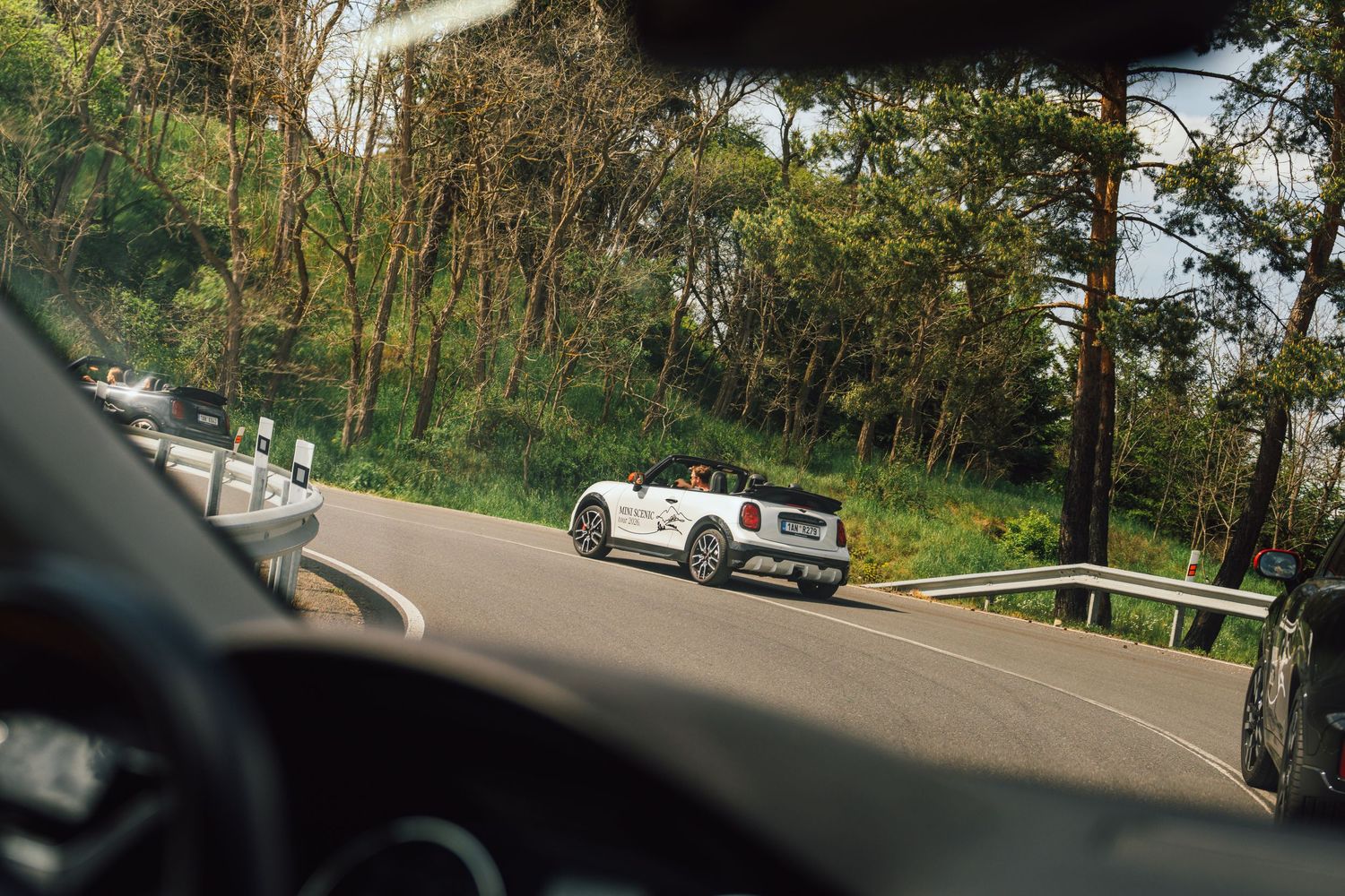 White MINI Cooper convertible photographed from inside a following car on a winding forest road.