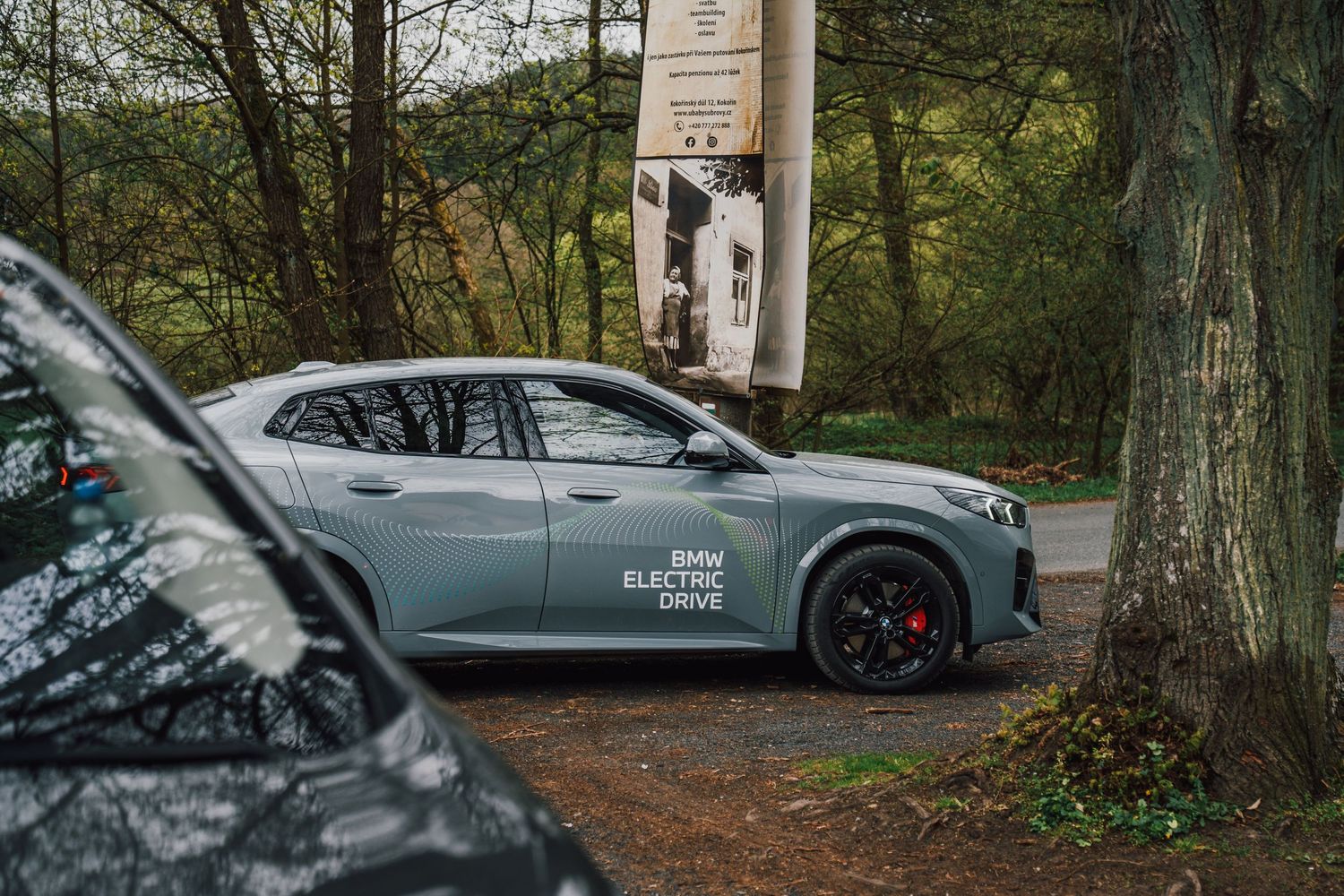 A grey BMW Electric Drive SUV parked near trees beside an information board in a forest setting.