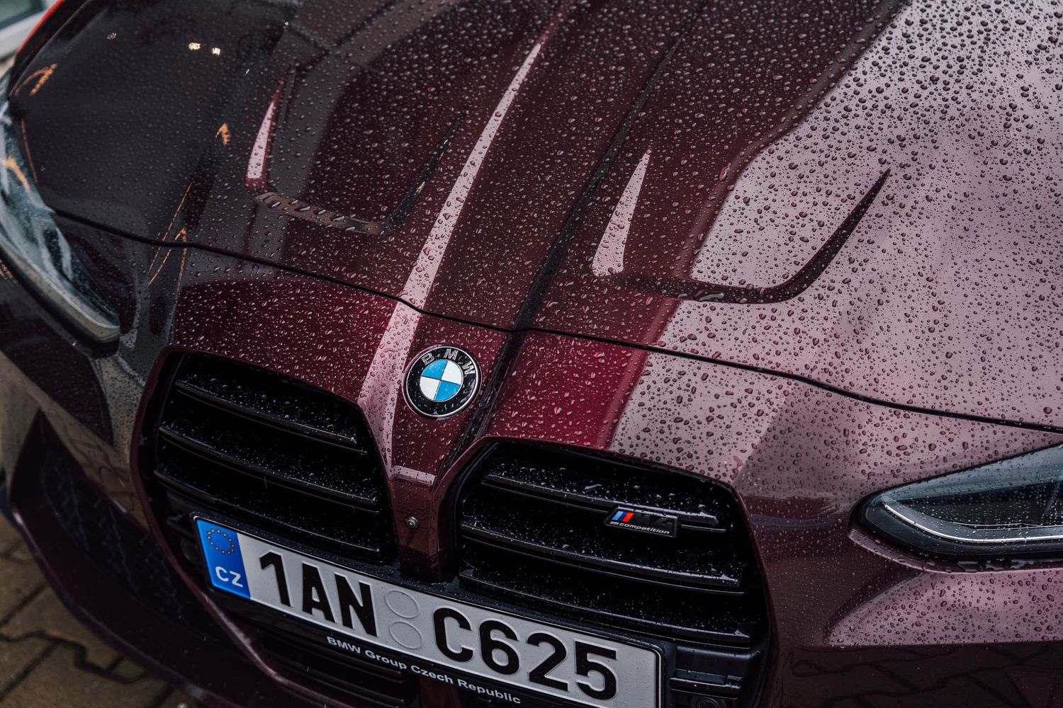 Close-up of a rain-covered dark red BMW M4 hood featuring the iconic BMW roundel badge.