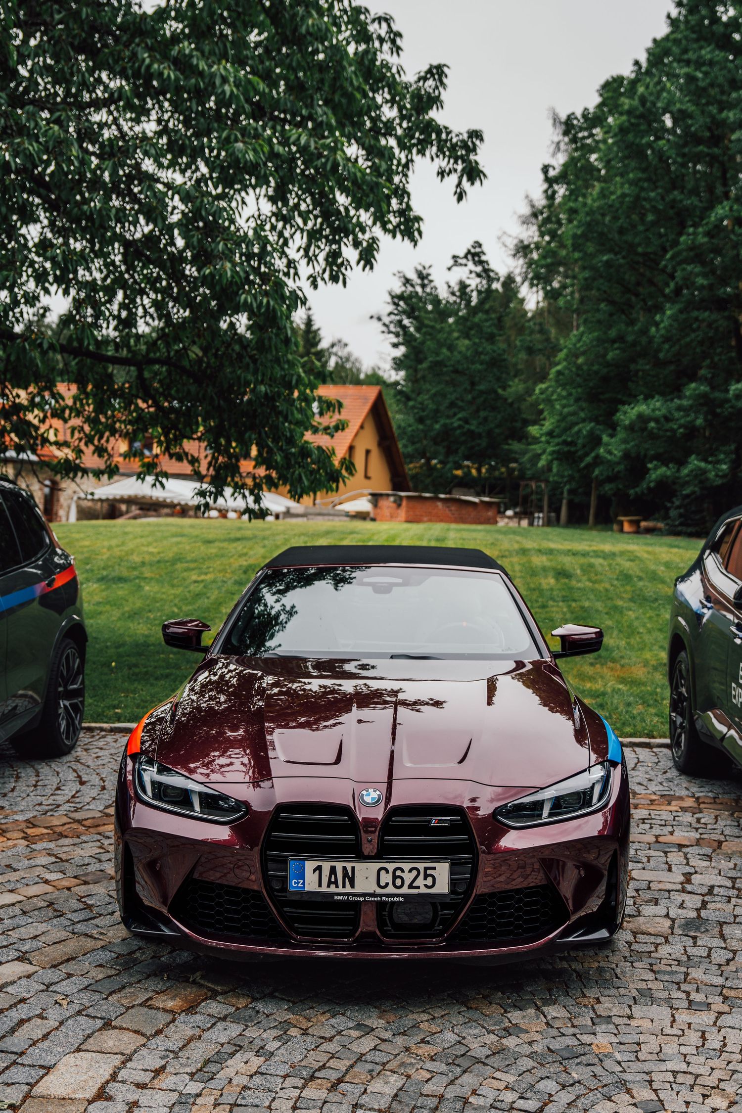 Stunning dark red BMW M4 parked on cobblestones surrounded by lush green trees.