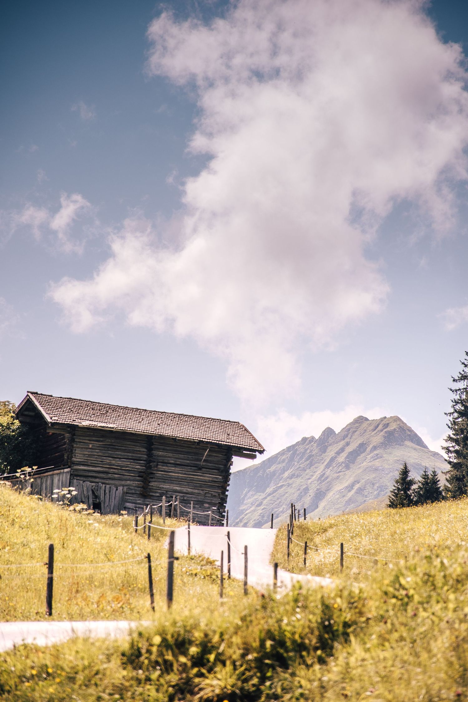 Old wooden alpine barn on green hillside with mountain peaks and dramatic cloudy sky.
