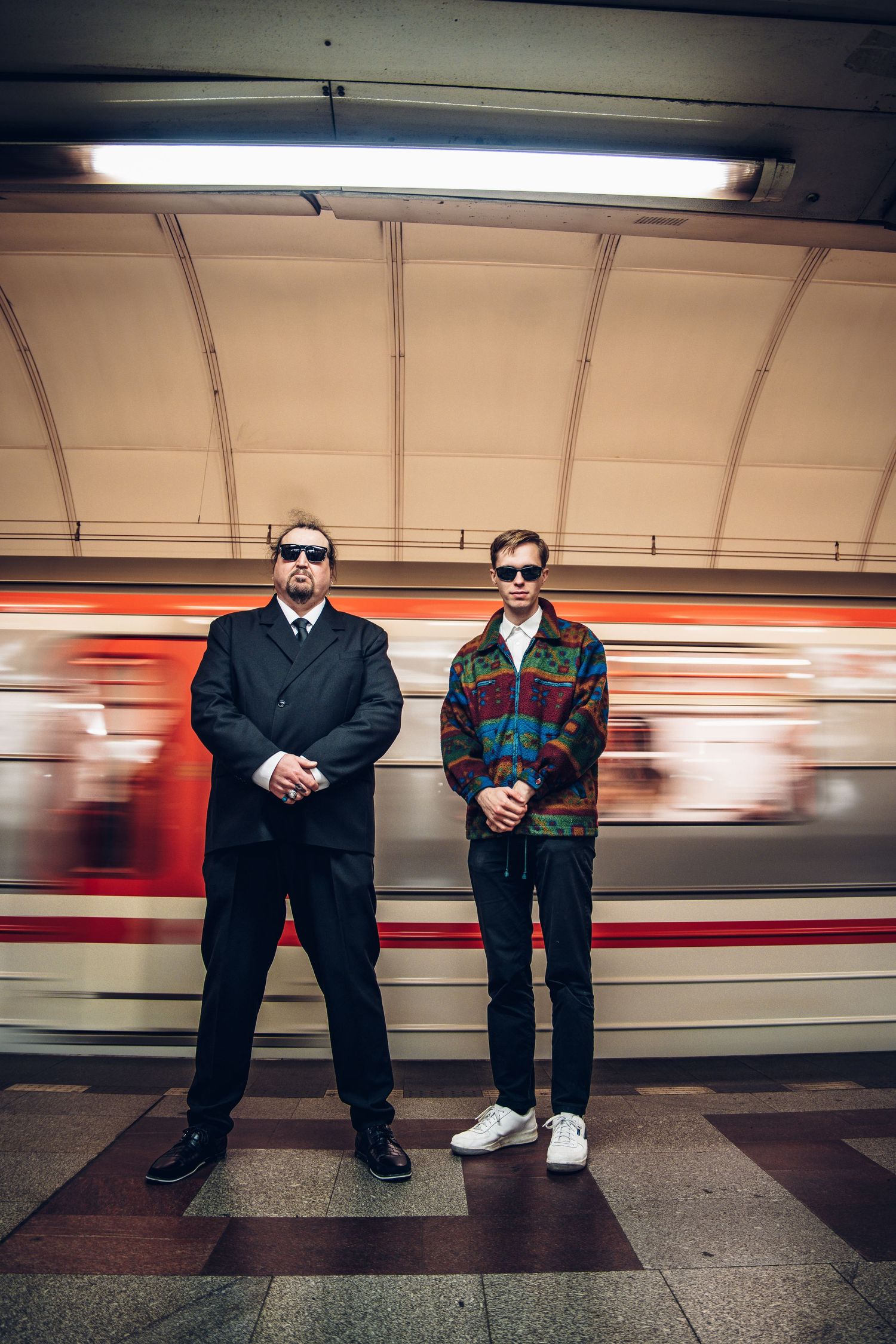Two men in sunglasses stand on a subway platform with a blurred passing train.