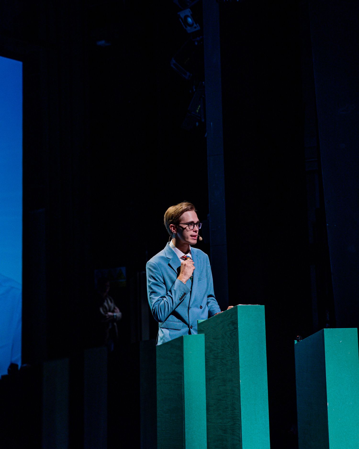 Young man in light blue suit speaking at podium on stage with teal green panels during conference event.